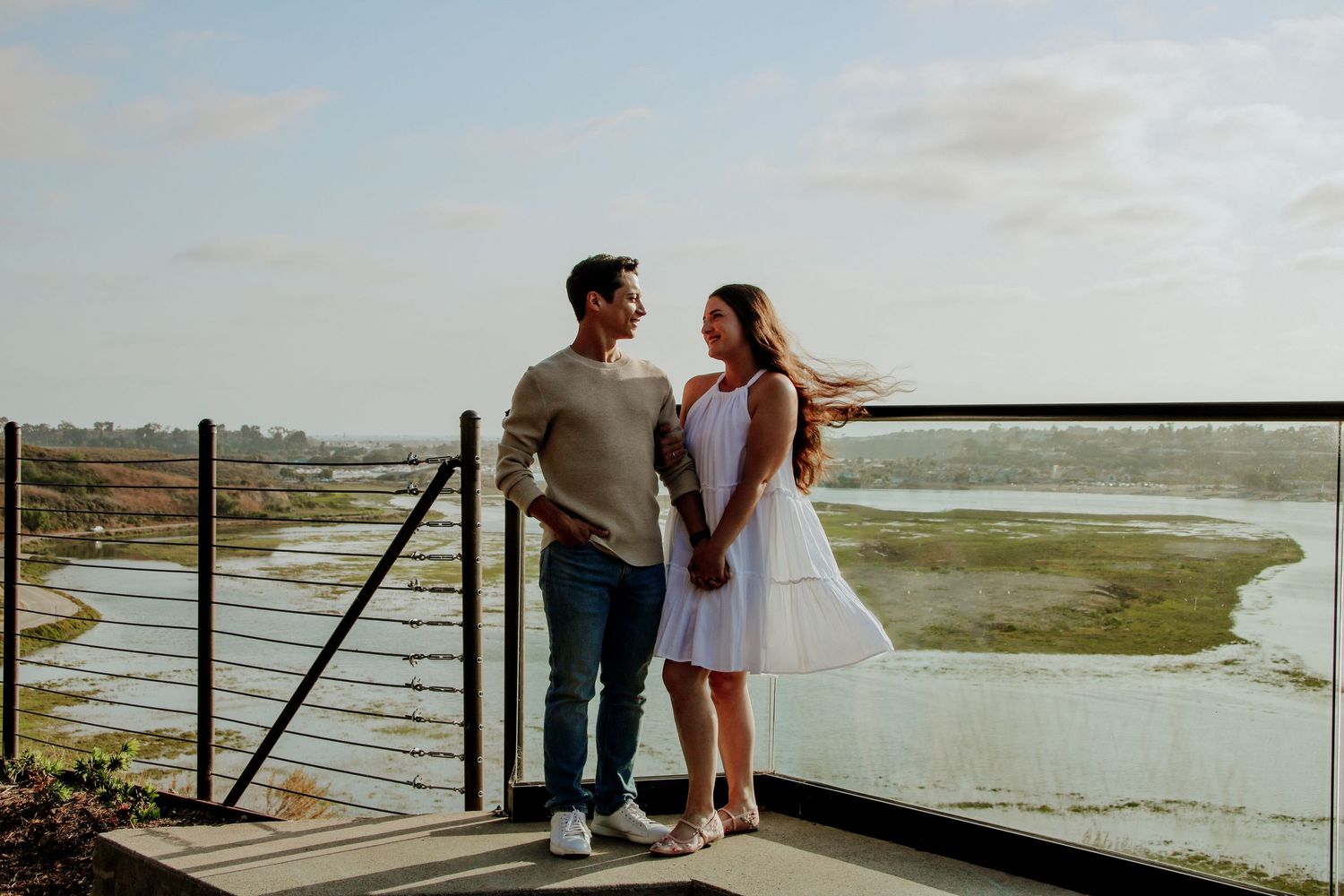 A couple stands close together on a wooden deck overlooking marshland during golden hour.