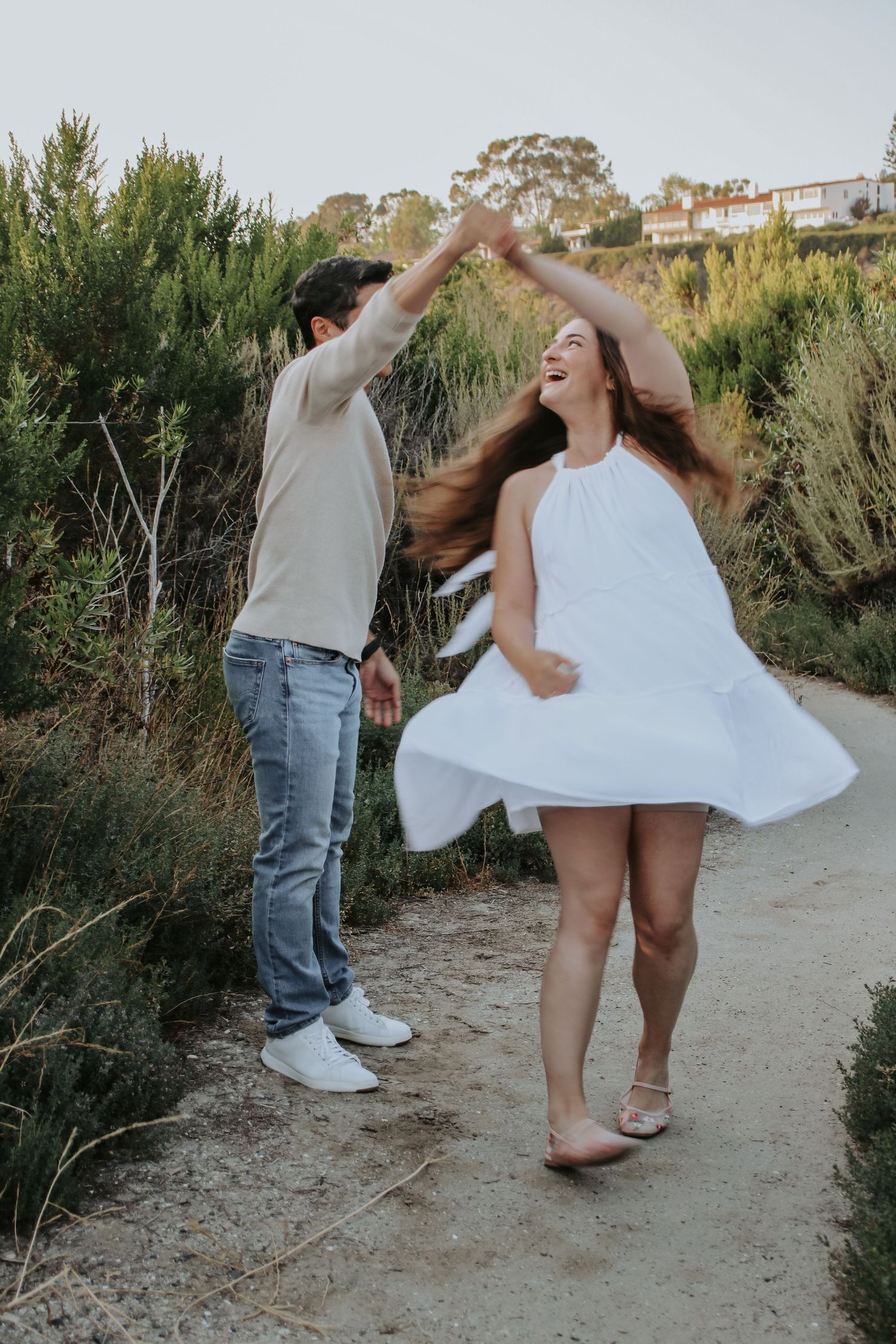 A couple dances joyfully on a coastal trail with the woman's white dress flowing in the movement.