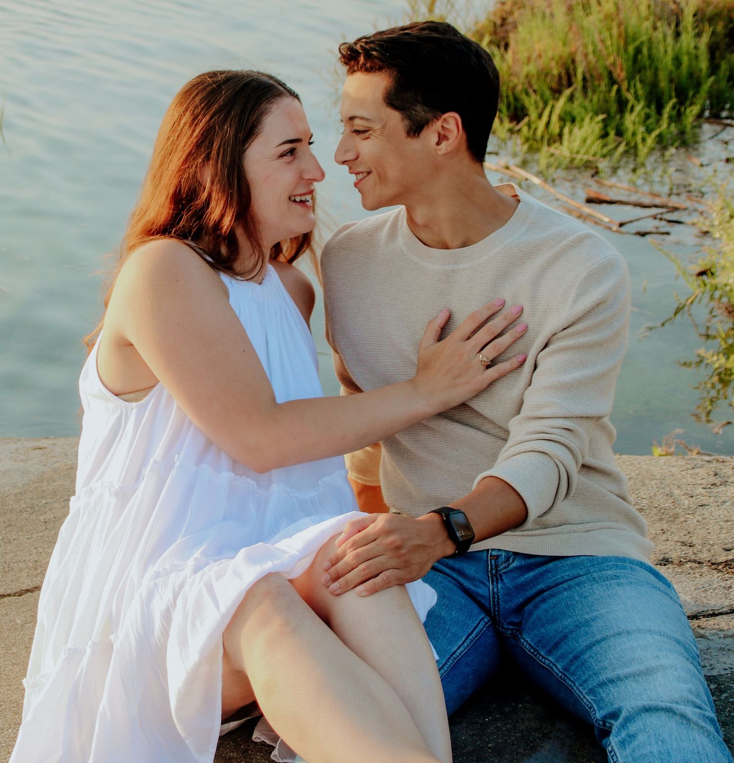 Couple wearing white and beige embracing on rocky shoreline with water in background.