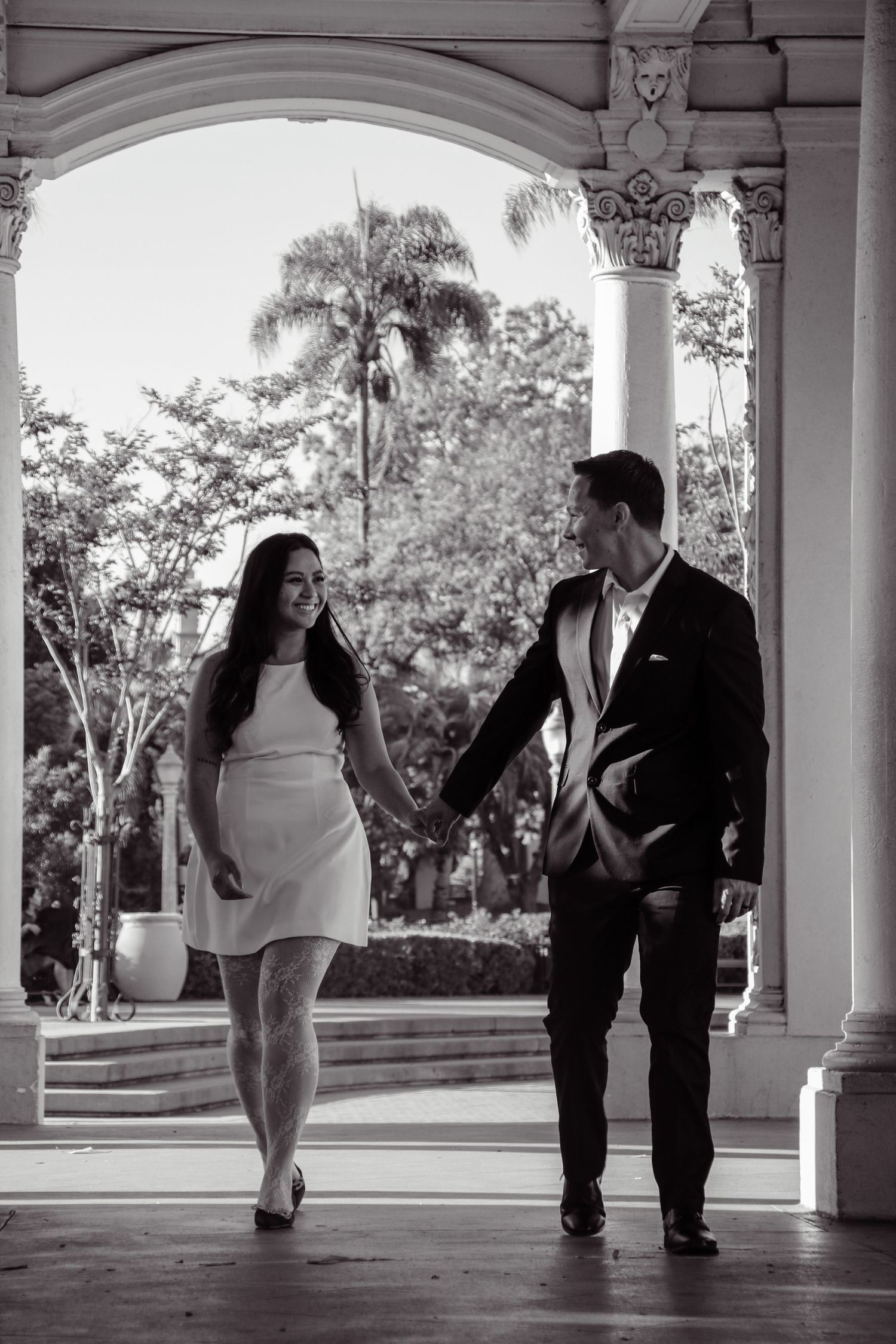 Black and white photo of a couple walking together under elegant architectural arches.