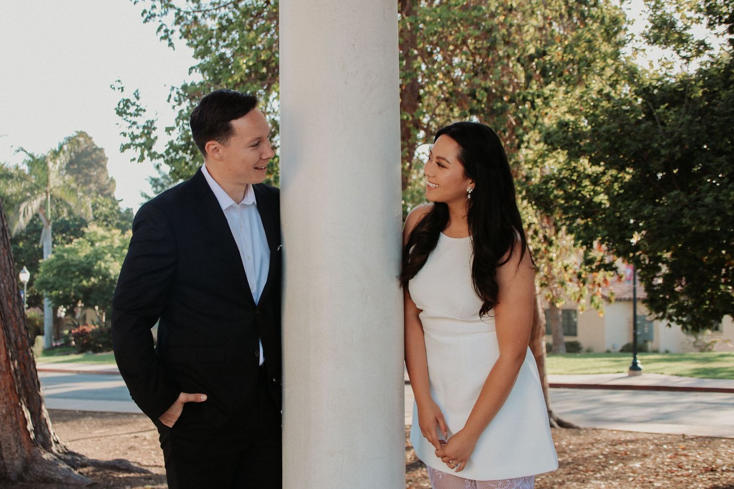 A couple in formal attire poses against a white pillar in a park setting.