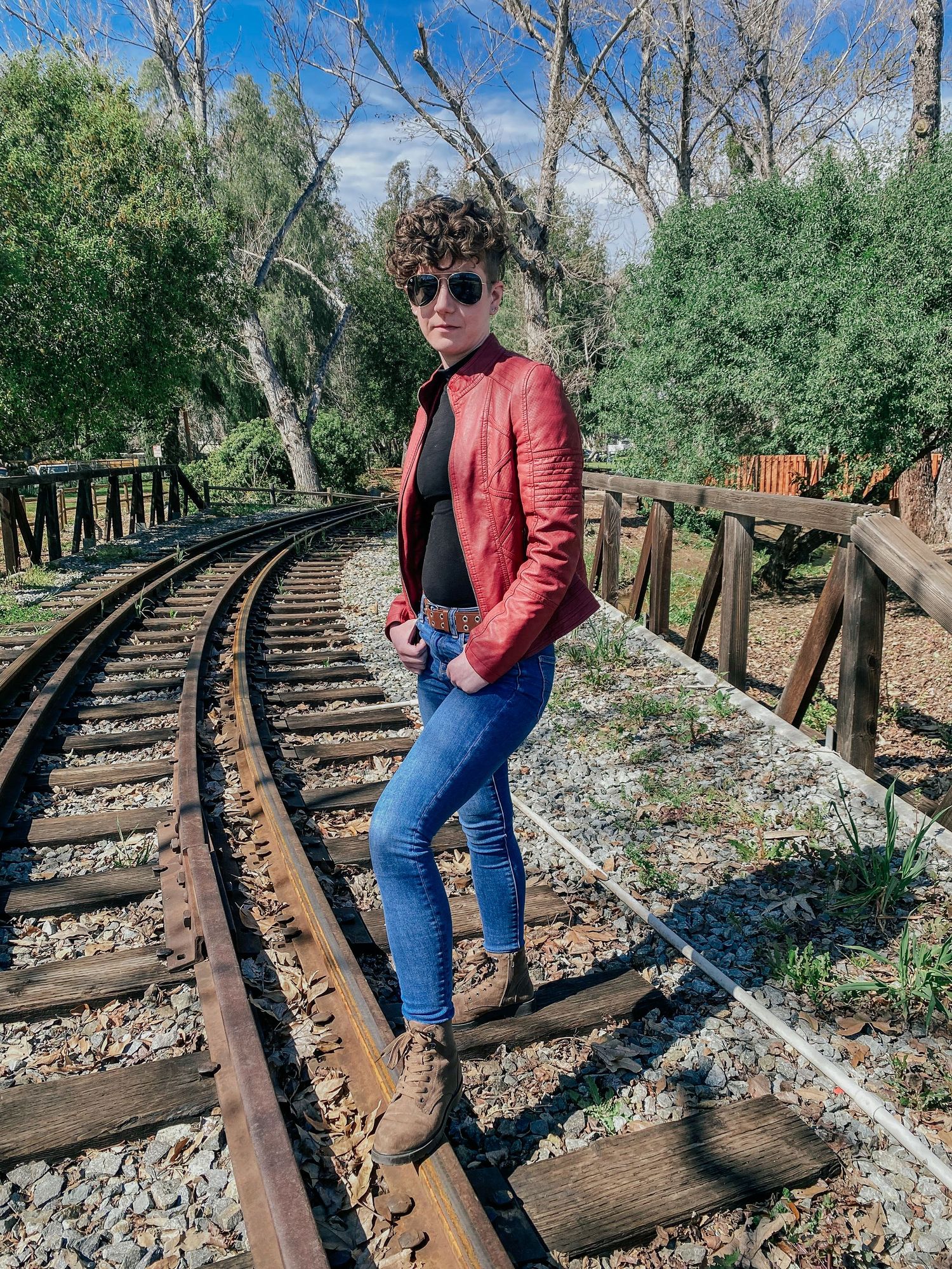 Railroad track photoshoot showcasing red leather jacket and sunglasses in natural outdoor setting.