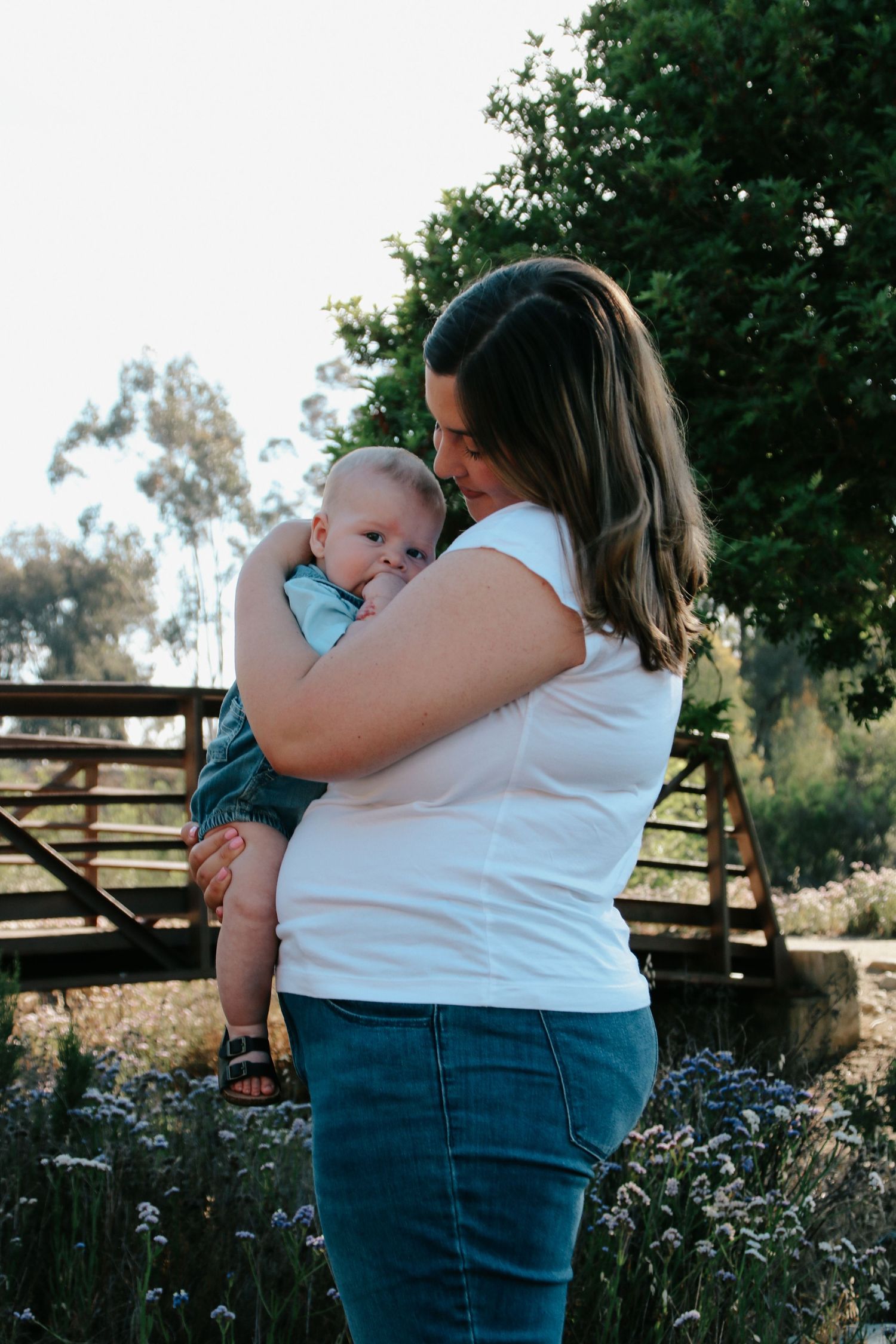 Mother holding baby against natural outdoor setting with wooden fence and greenery.