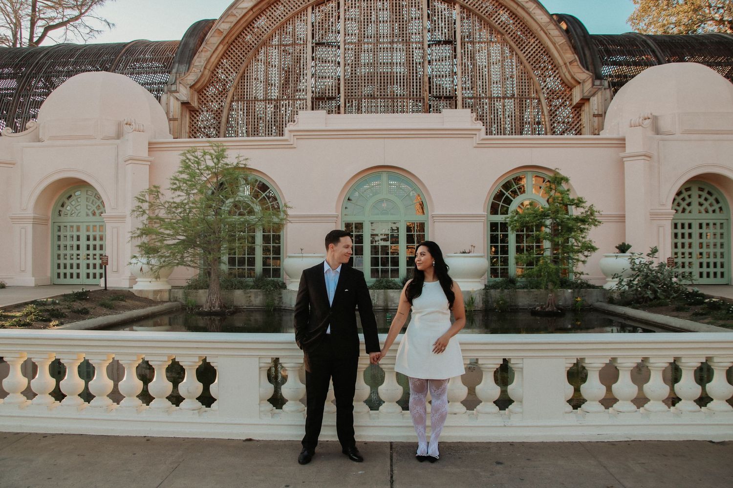 Elegant couple standing before grand archway and stone balustrade of historic building.