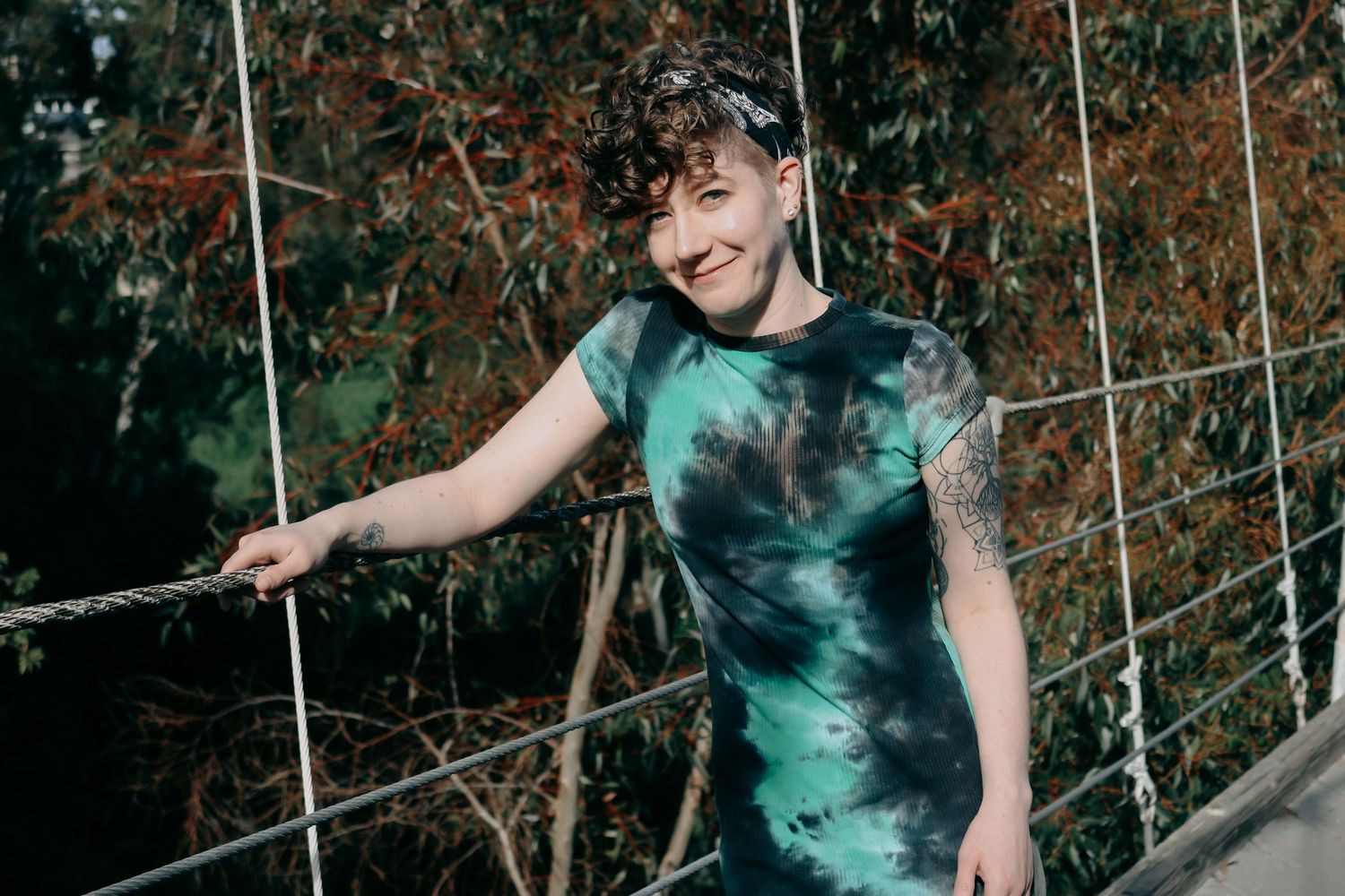 A person in a tie-dye green shirt poses energetically on a wooden bridge with metal cables.