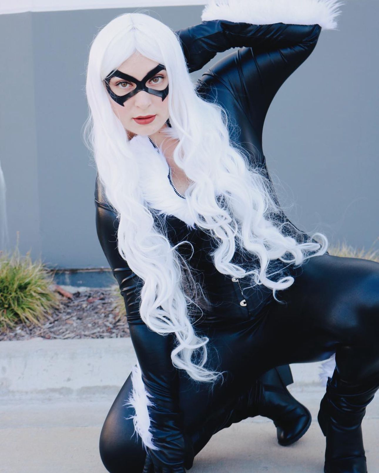 A cosplayer in a black catsuit and white wig strikes a dynamic pose against a gray wall.