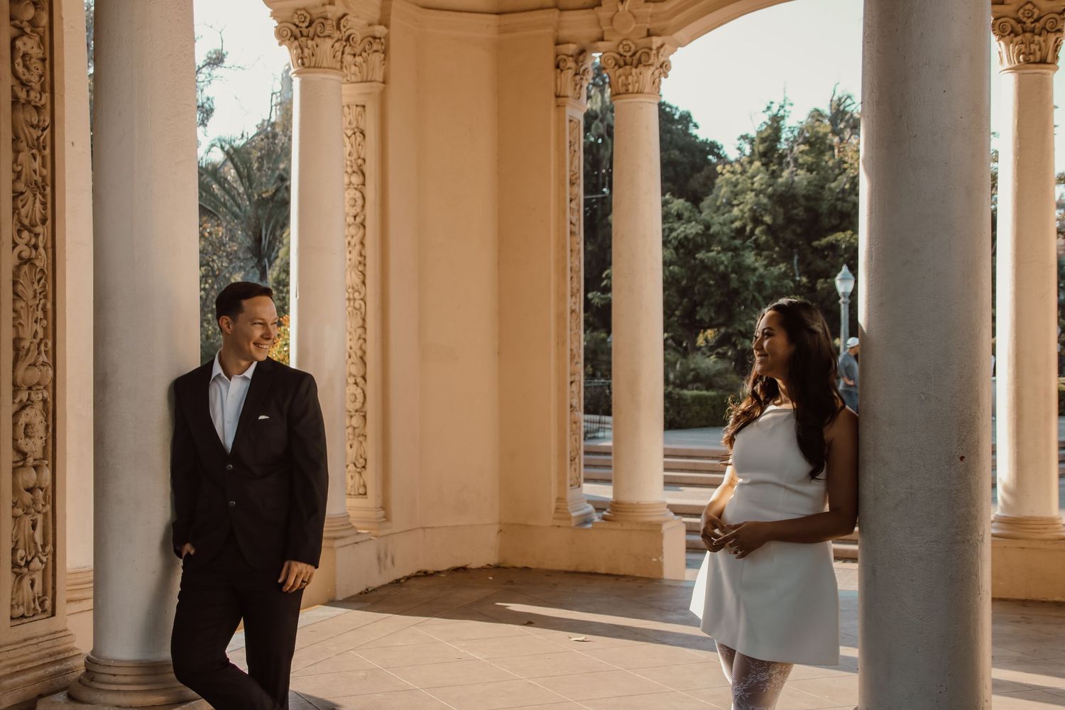 A couple poses in a sunlit classical colonnade with ornate Corinthian columns.