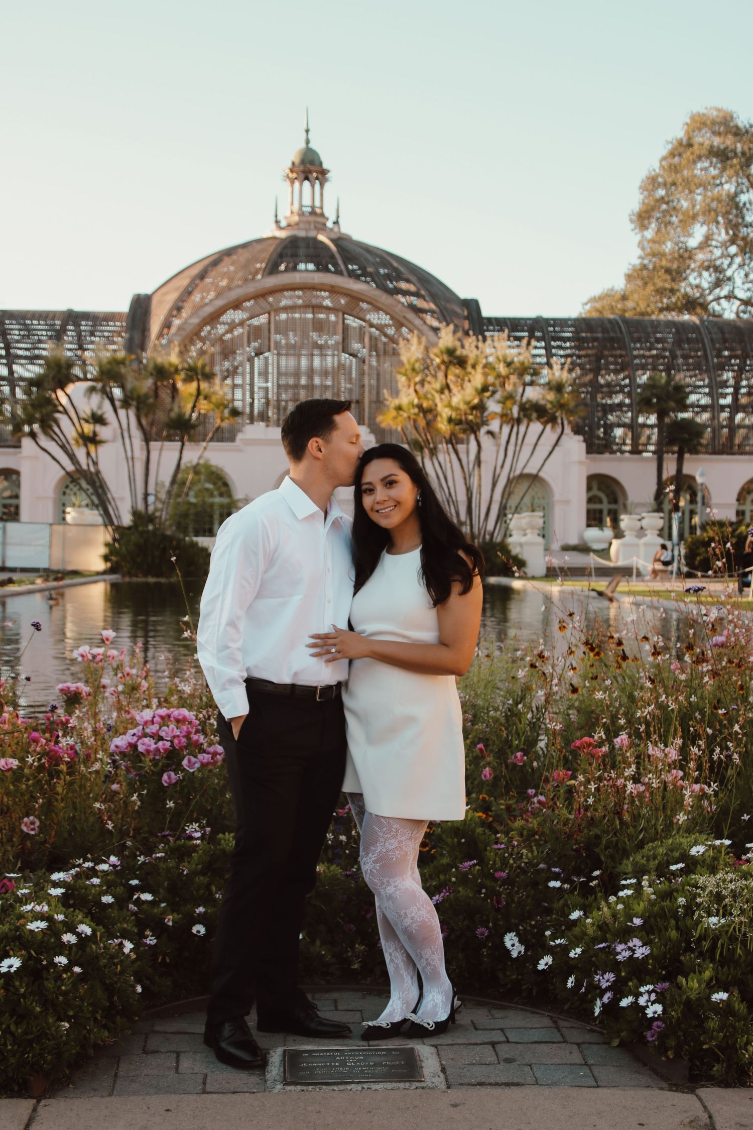 A couple shares loving moments in a garden surrounded by colorful flowers and elegant architecture.