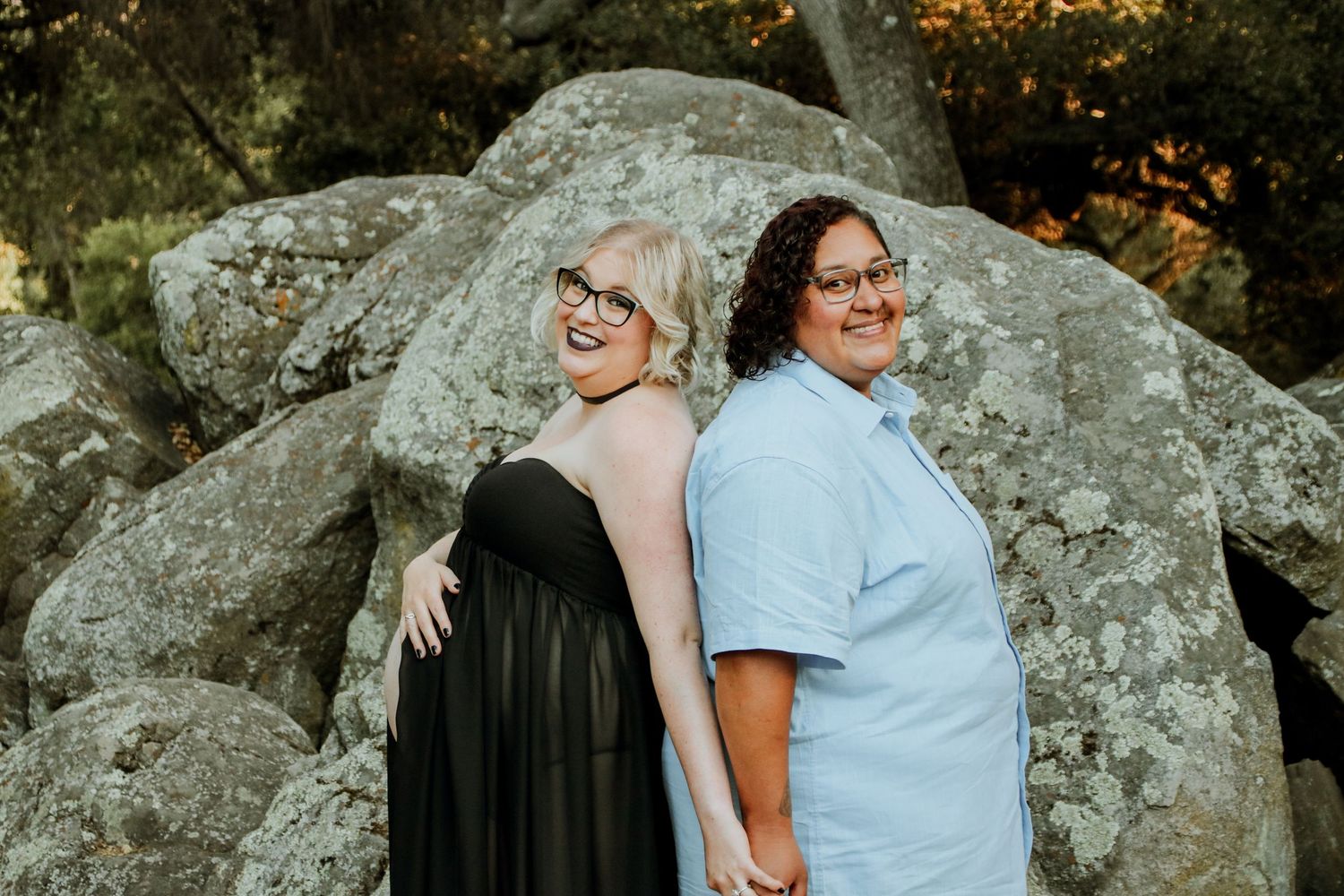 Two people lean back-to-back against a large boulder while posing for a maternity photo.