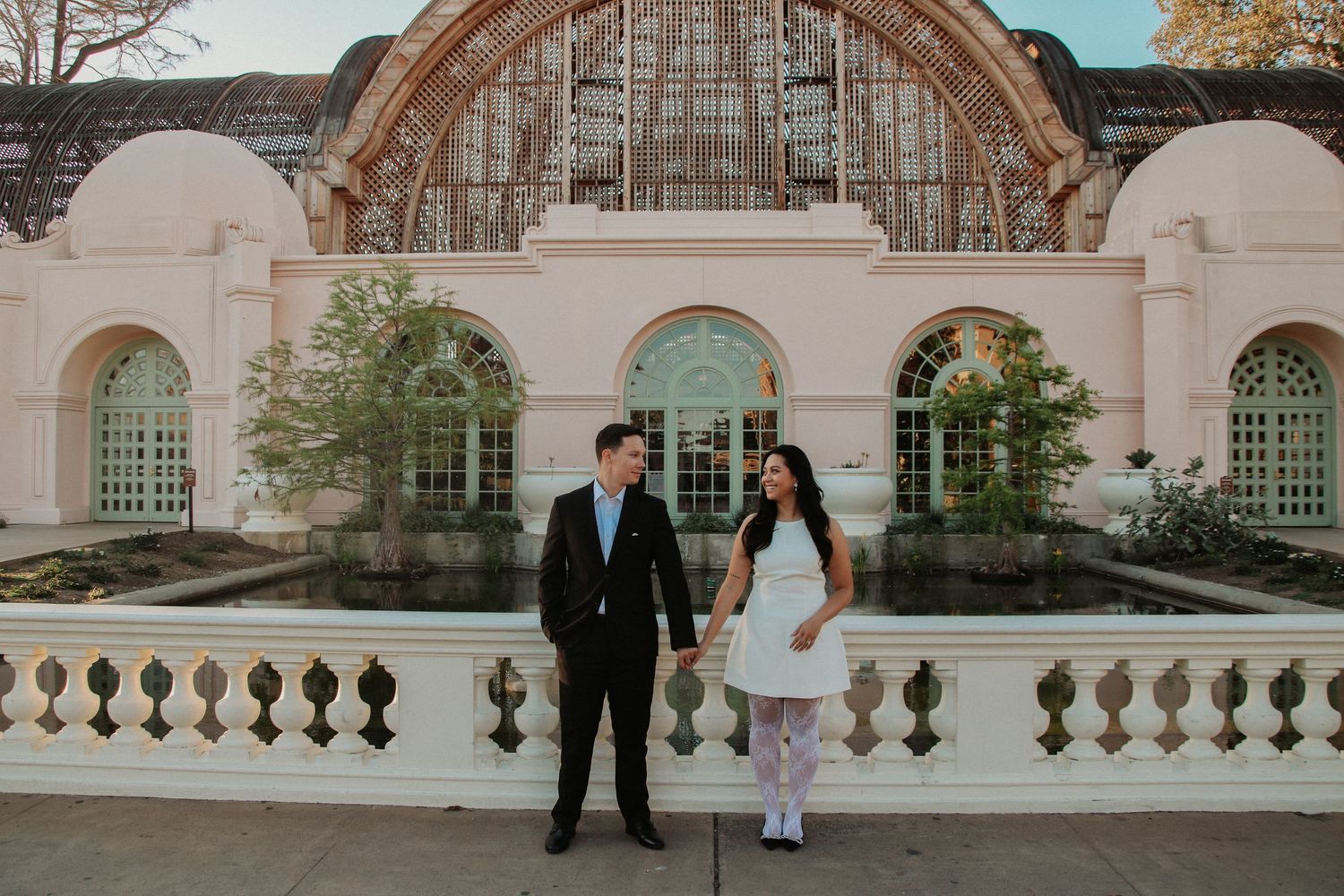 Couple posing in front of ornate white arched building with elegant balustrade and architectural details.