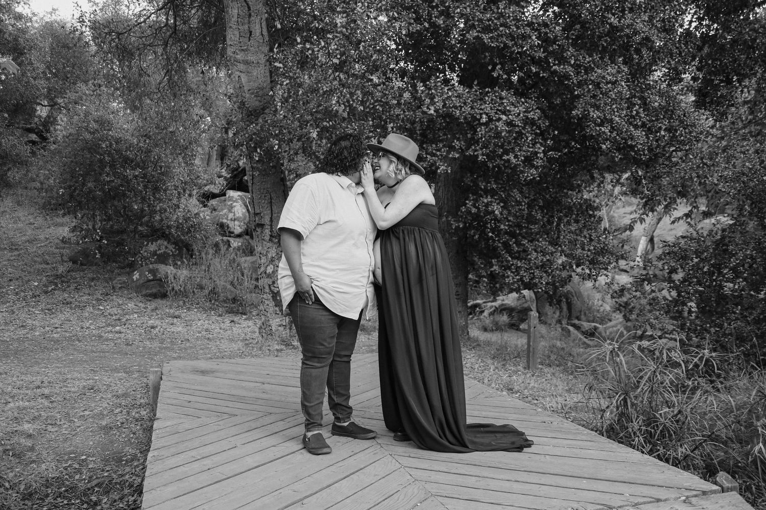 Black and white photo of couple standing together on wooden deck surrounded by nature.