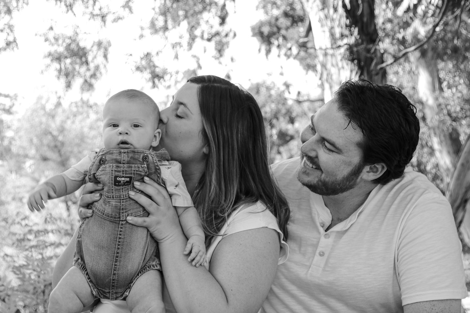 A tender family moment captured in black and white as parents share affection with their baby in an outdoor setting.