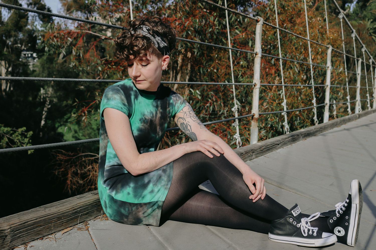 A person wearing a green tie-dye outfit relaxes on a bridge deck with autumn trees in background.