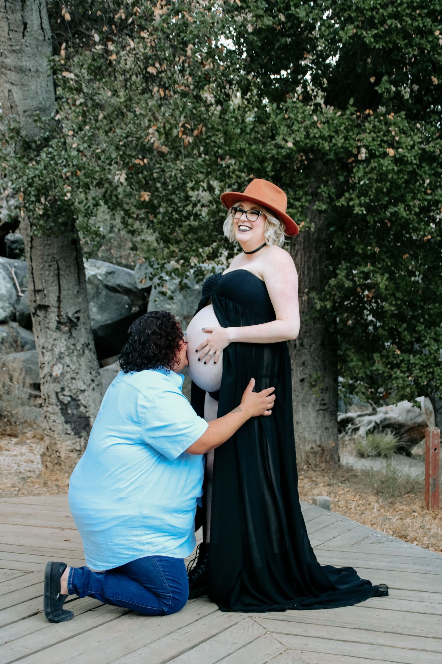 A maternity photoshoot outdoors showing a couple on a wooden deck with trees in the background.