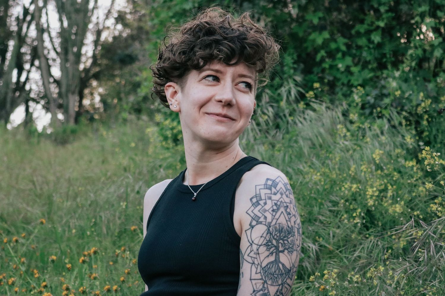 Person with sleeve tattoo sits in black dress against natural backdrop.