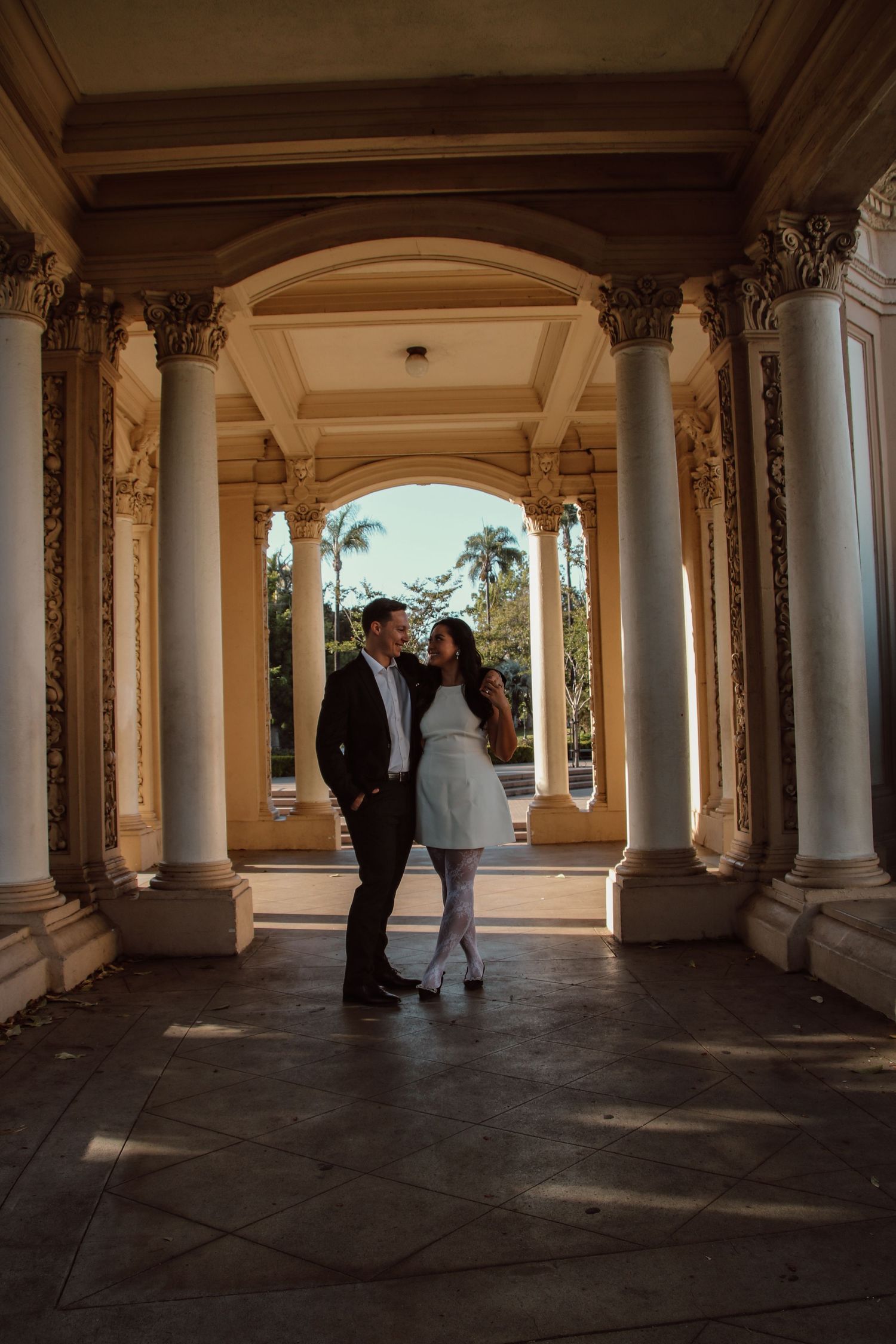 Two people stand close together in a grand columned walkway with beautiful golden hour lighting.