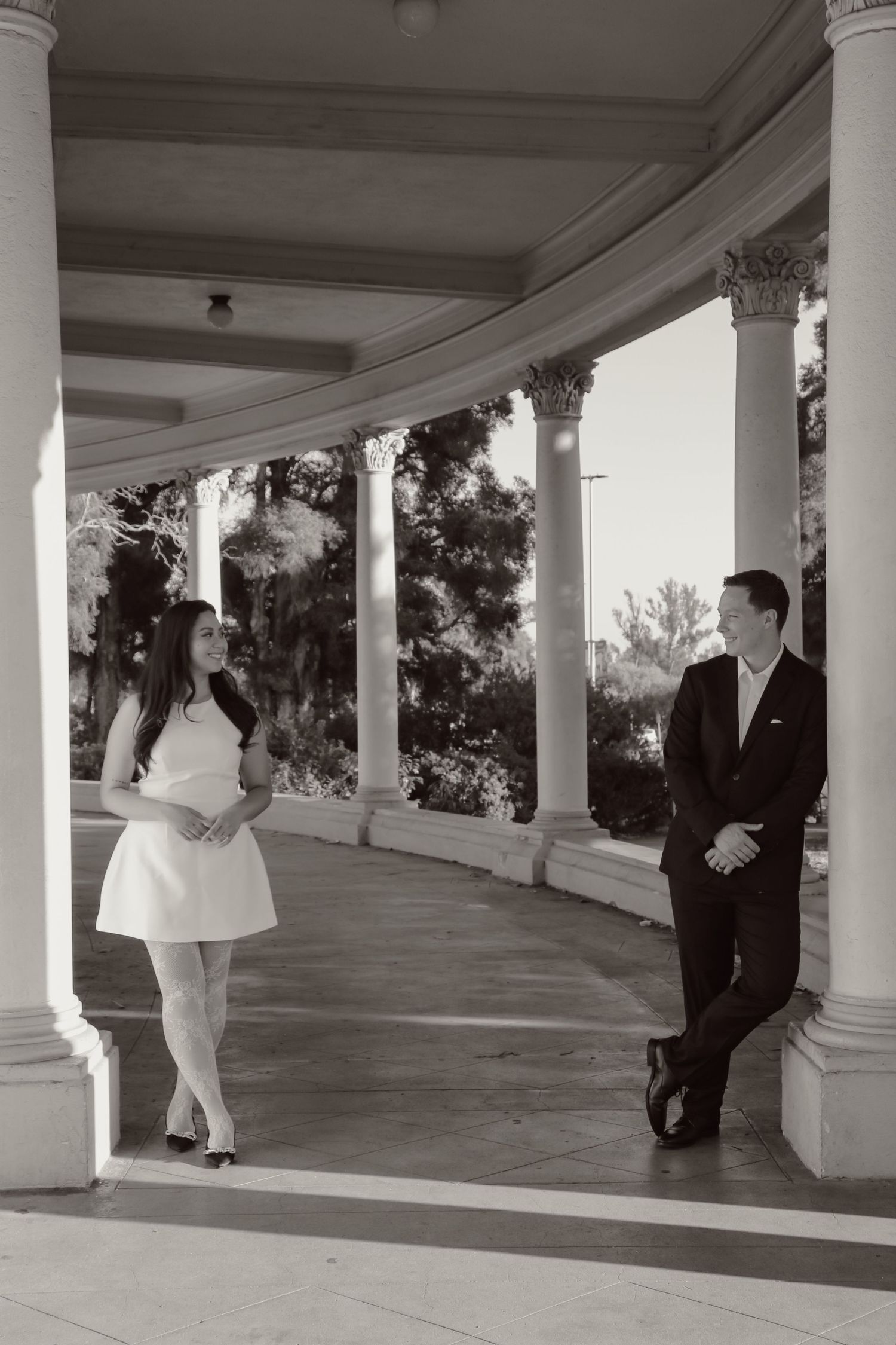A couple in formal attire poses in black and white along a columned walkway.
