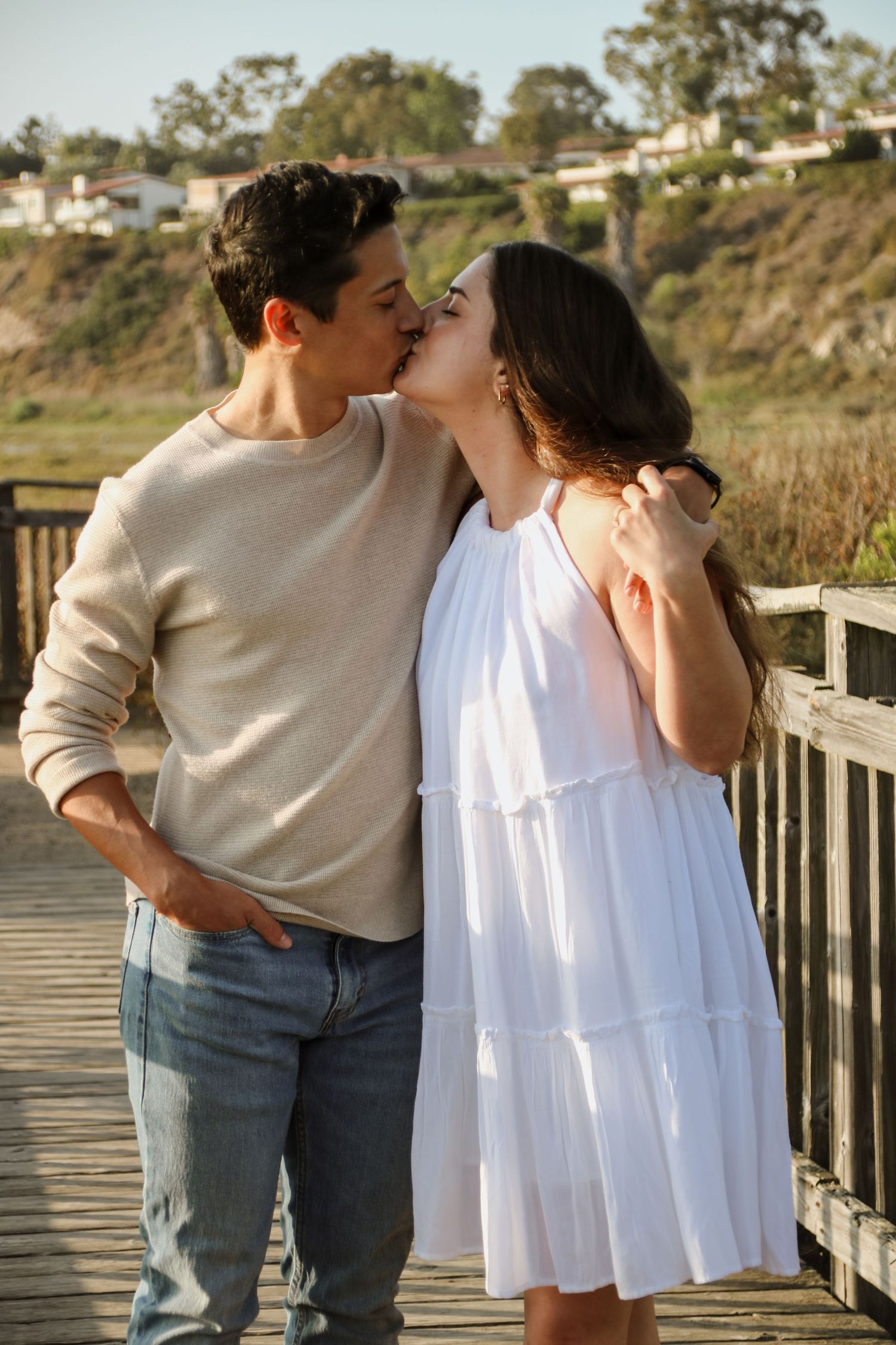 A romantic couple sharing a tender kiss on a wooden boardwalk with coastal cliffs and houses in the background.