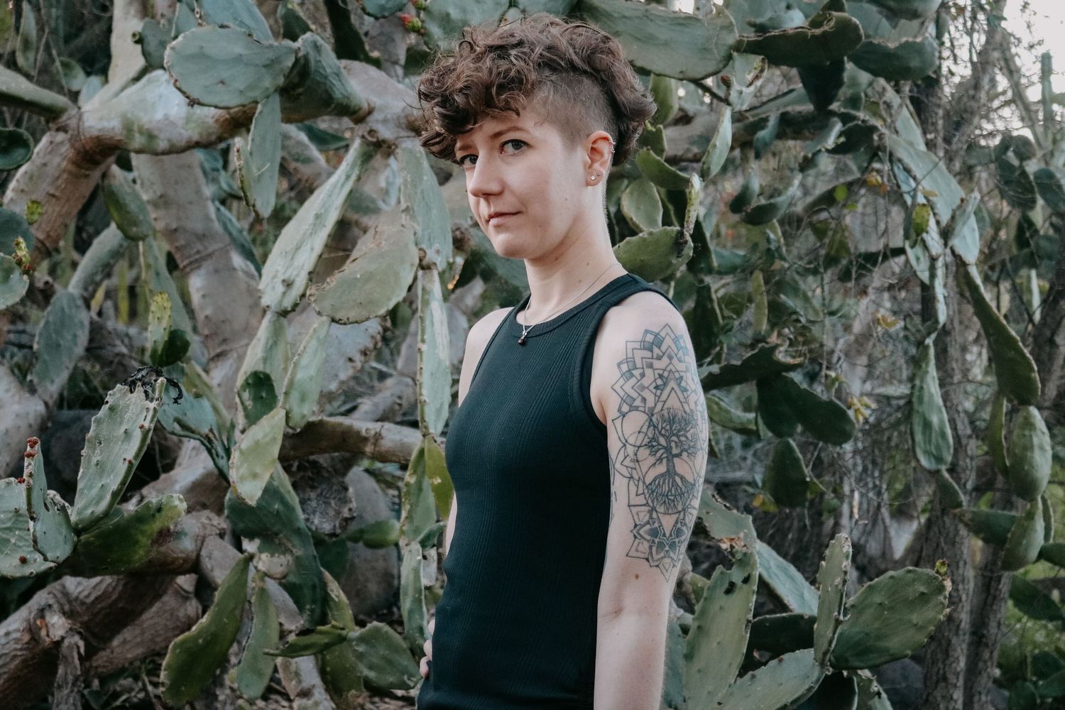 Modern portrait against desert plants featuring black sleeveless top and detailed arm tattoo.