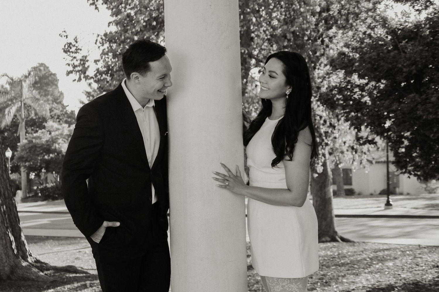A couple shares a playful moment while standing beside a white column in an outdoor setting.