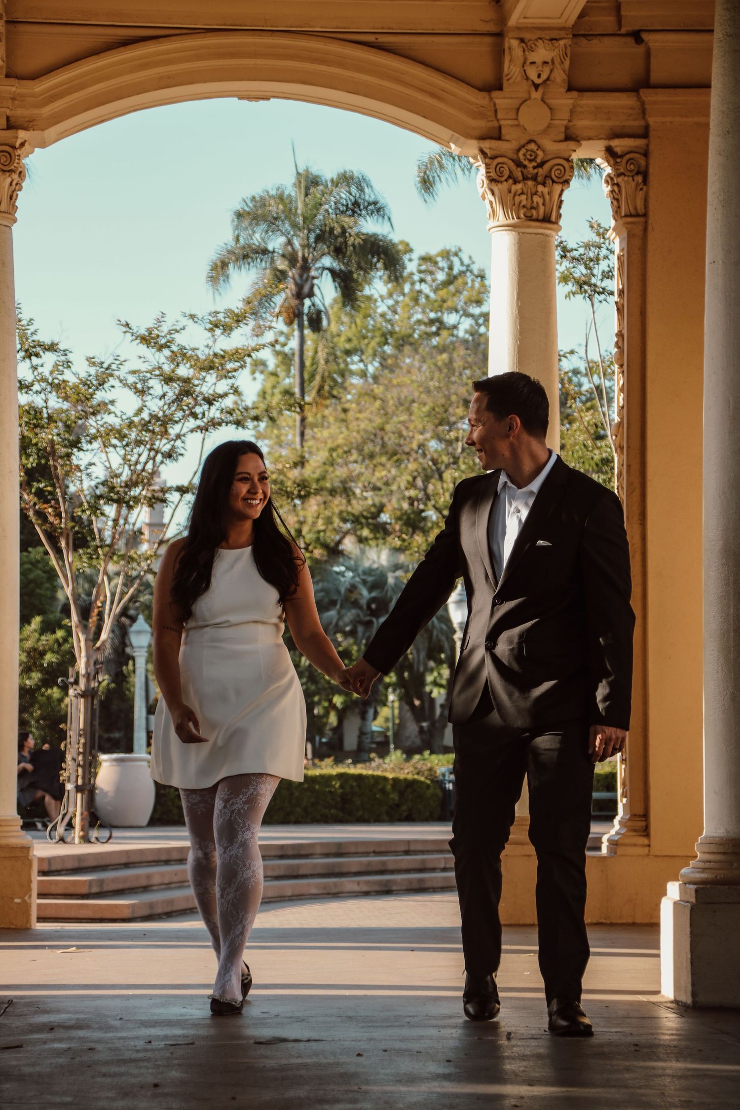 A couple shares a romantic moment while strolling through a colonnade with palm trees in background.