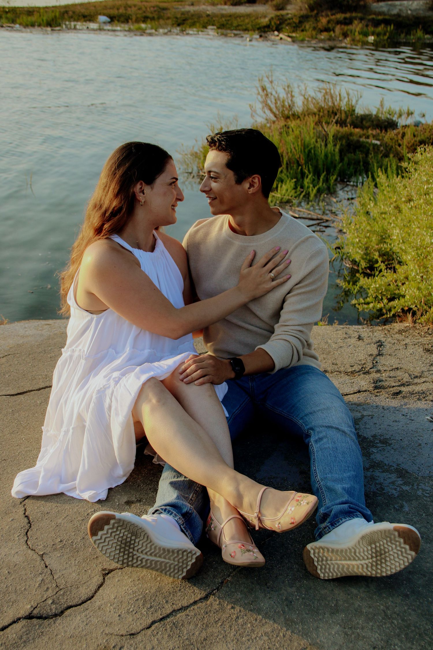 Couple in casual attire sharing a tender moment while sitting on rocks near a lake at sunset.