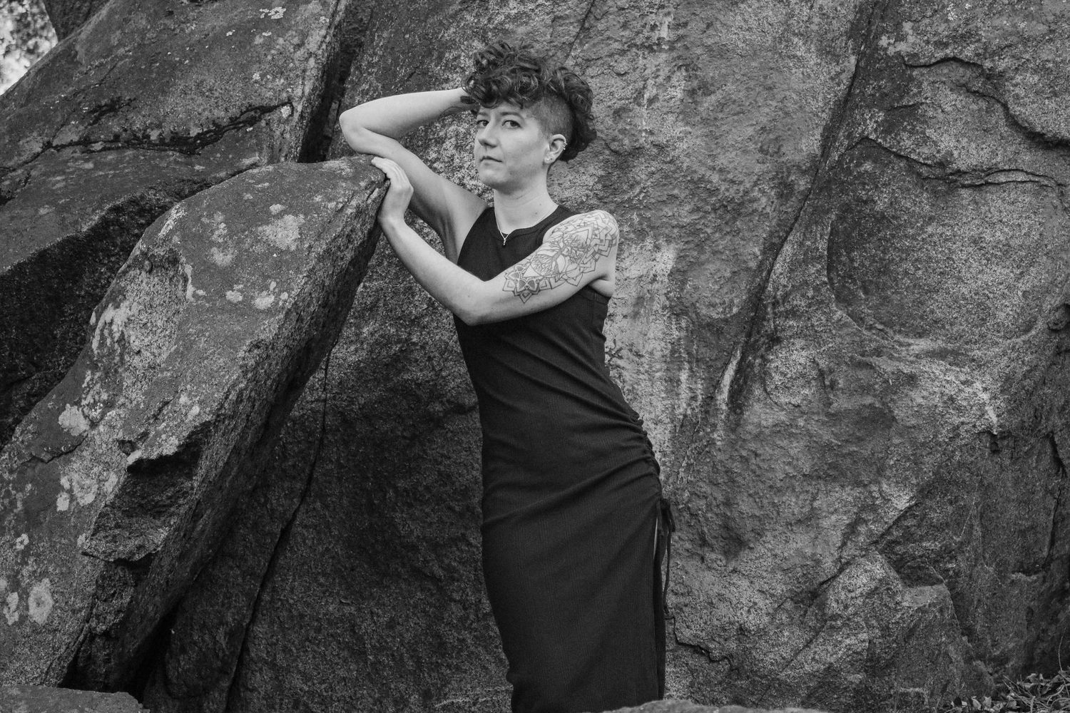 Dramatic monochrome photo of a person striking a pose against textured rock face wearing a black dress.