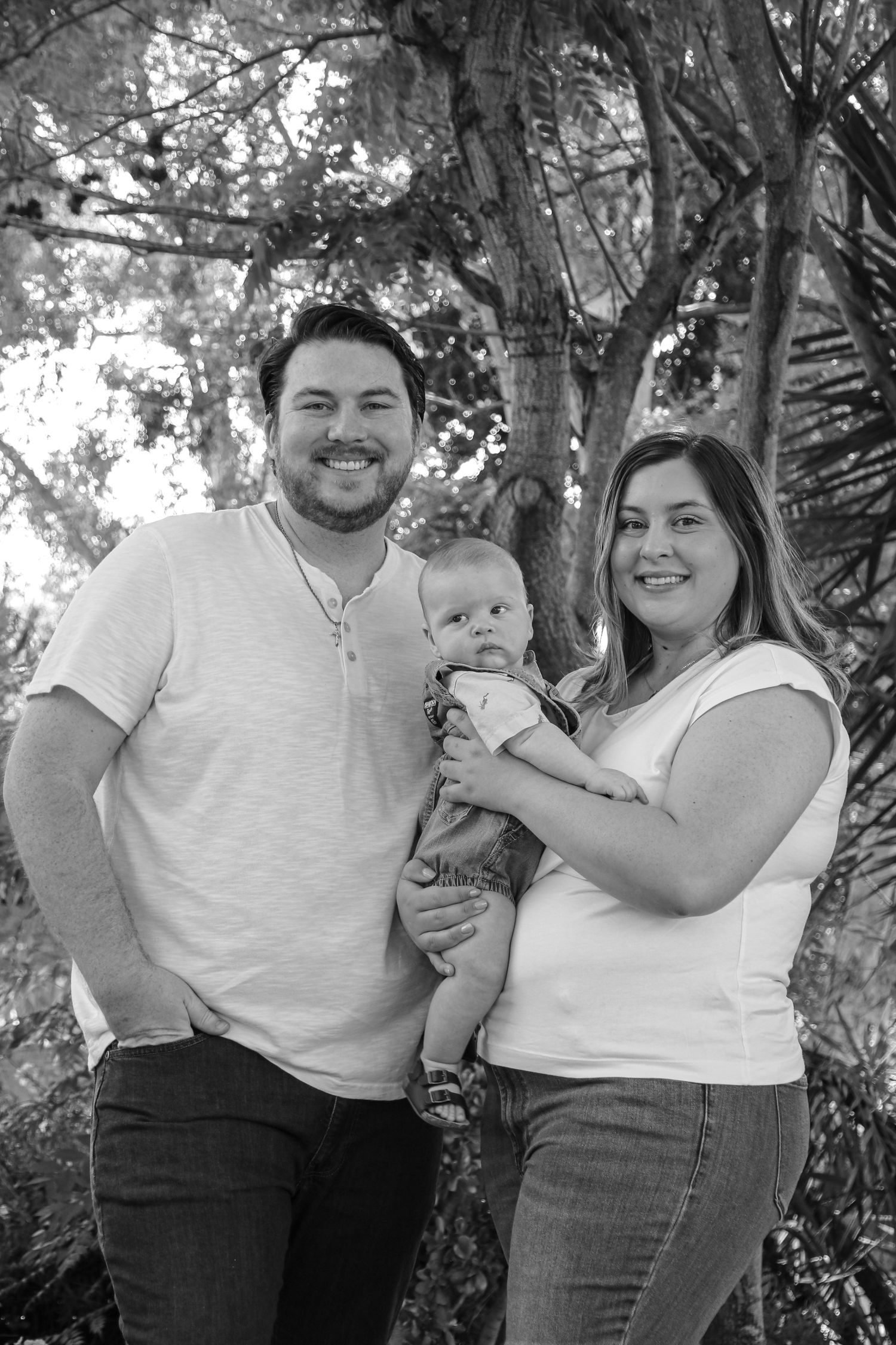 Black and white family portrait with parents holding baby against wooded background.