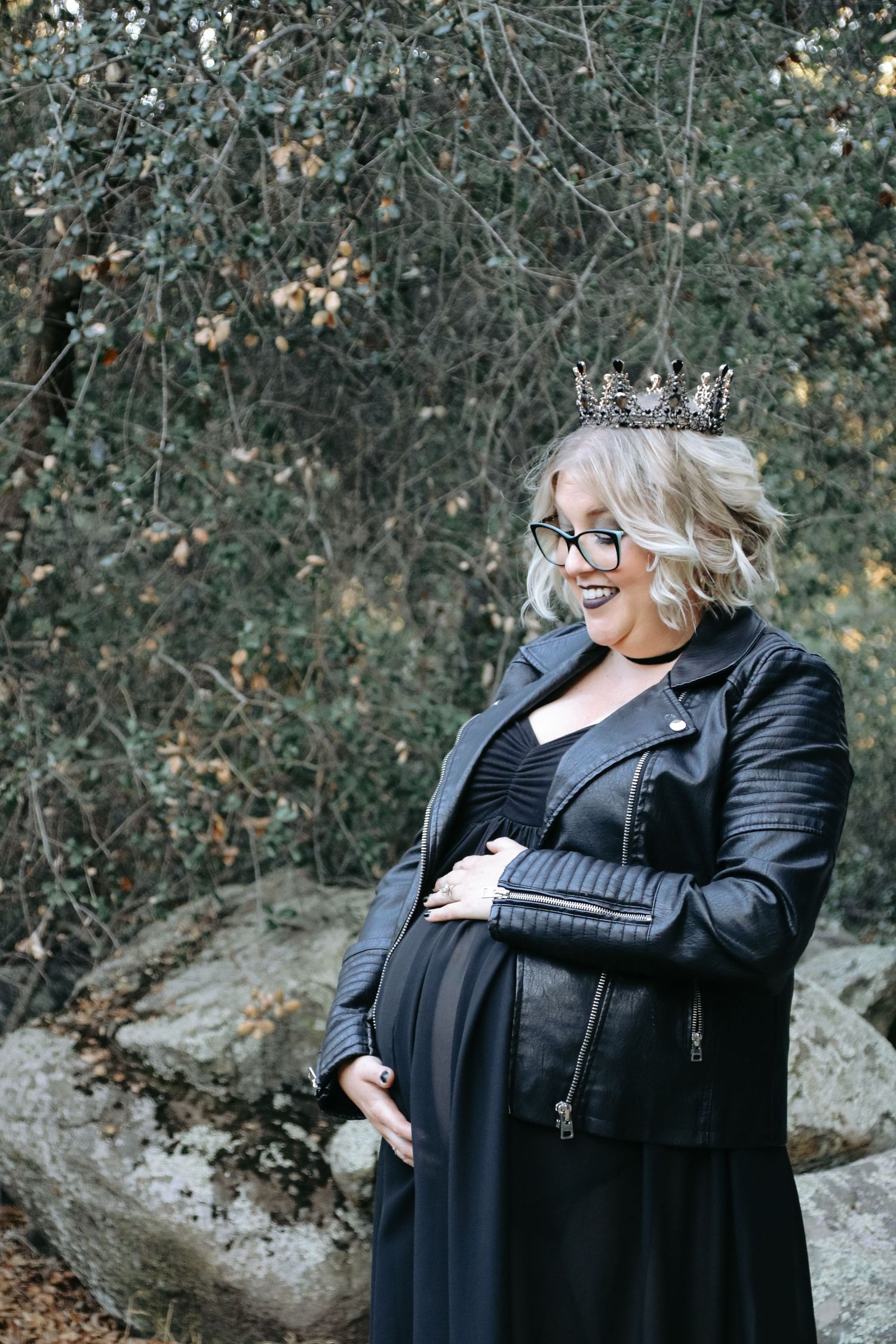 A maternity portrait with silver crown accessory, black leather jacket and dress against foliage background.