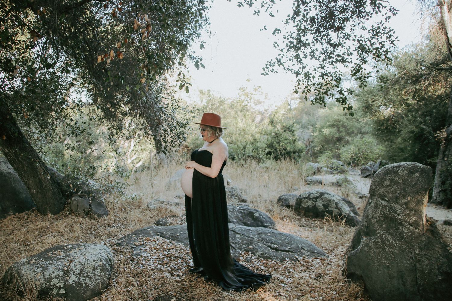 Figure in a long black dress and brown hat poses elegantly in a natural outdoor setting with rocks and trees.