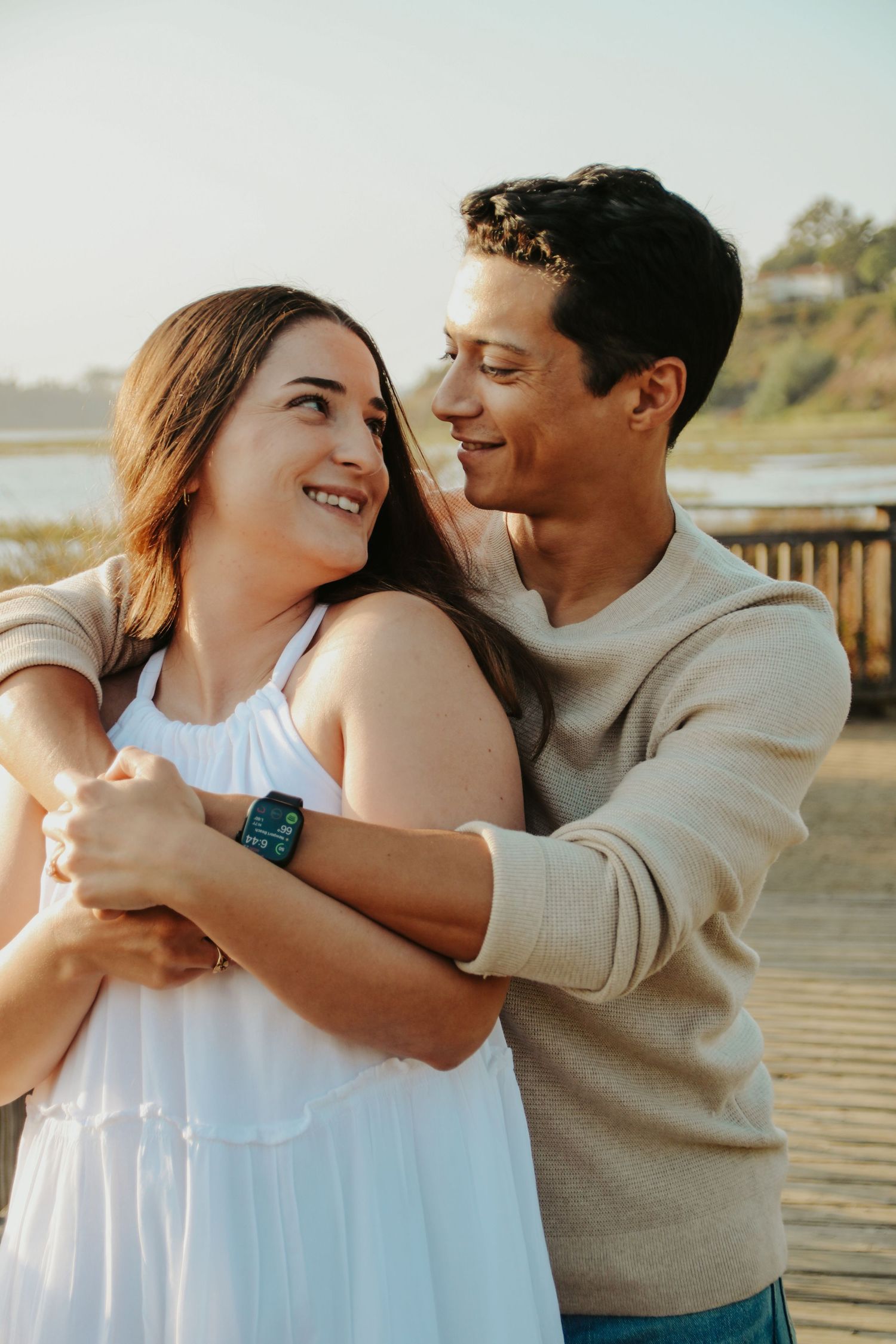 A couple shares a joyful embrace while laughing together on a seaside walkway at sunset.