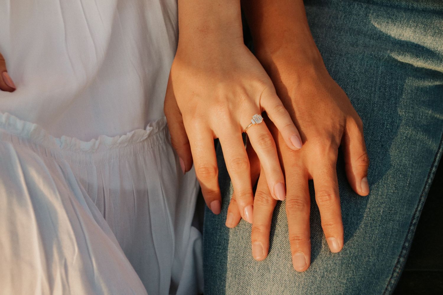 Close-up of hands with engagement rings resting together on white dress and denim.