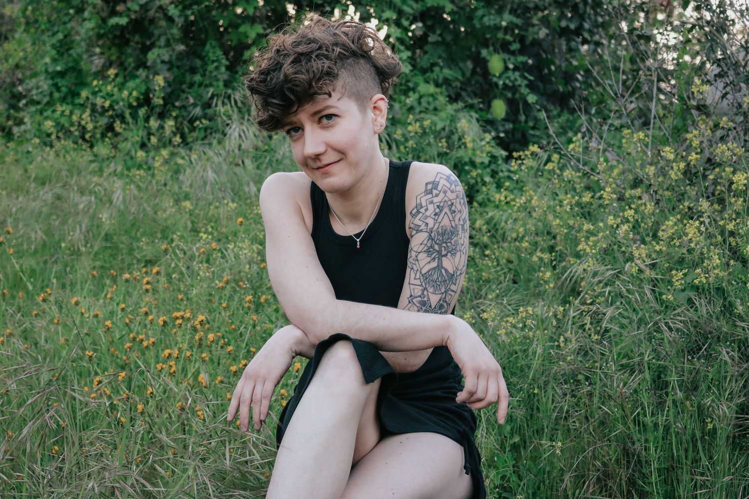 Model wearing black tank top sits casually in field of yellow wildflowers with visible arm tattoo.