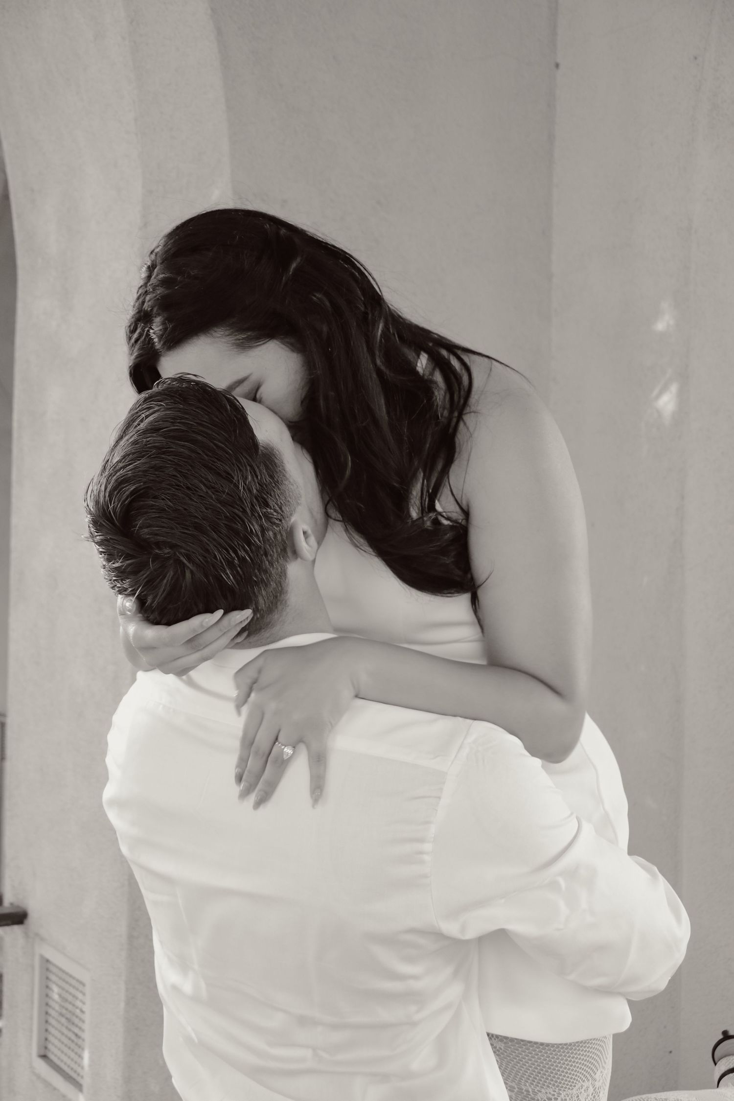 Black and white portrait of romantic couple embracing against neutral wall background.