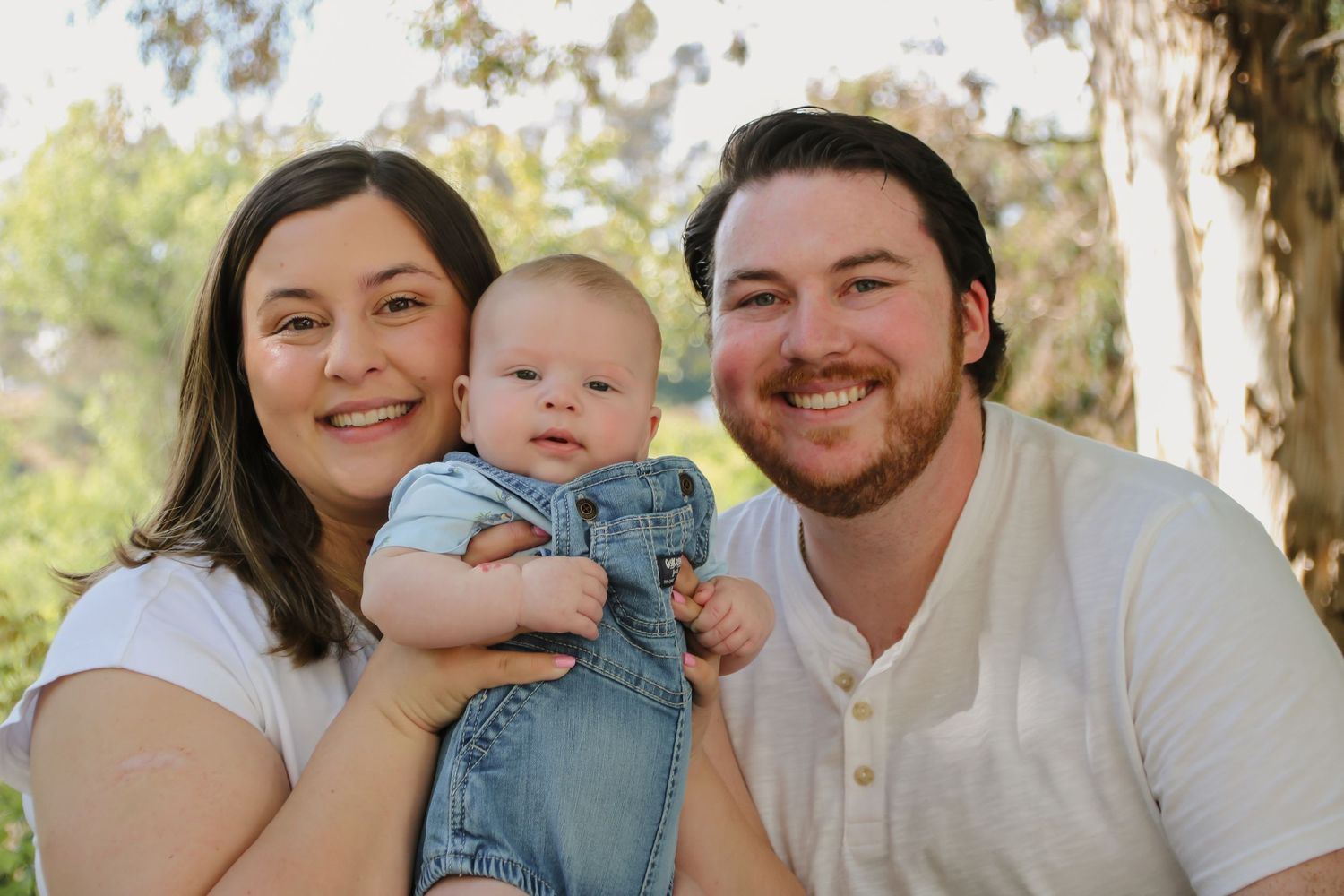 Family of three posing outdoors for portrait session under trees.