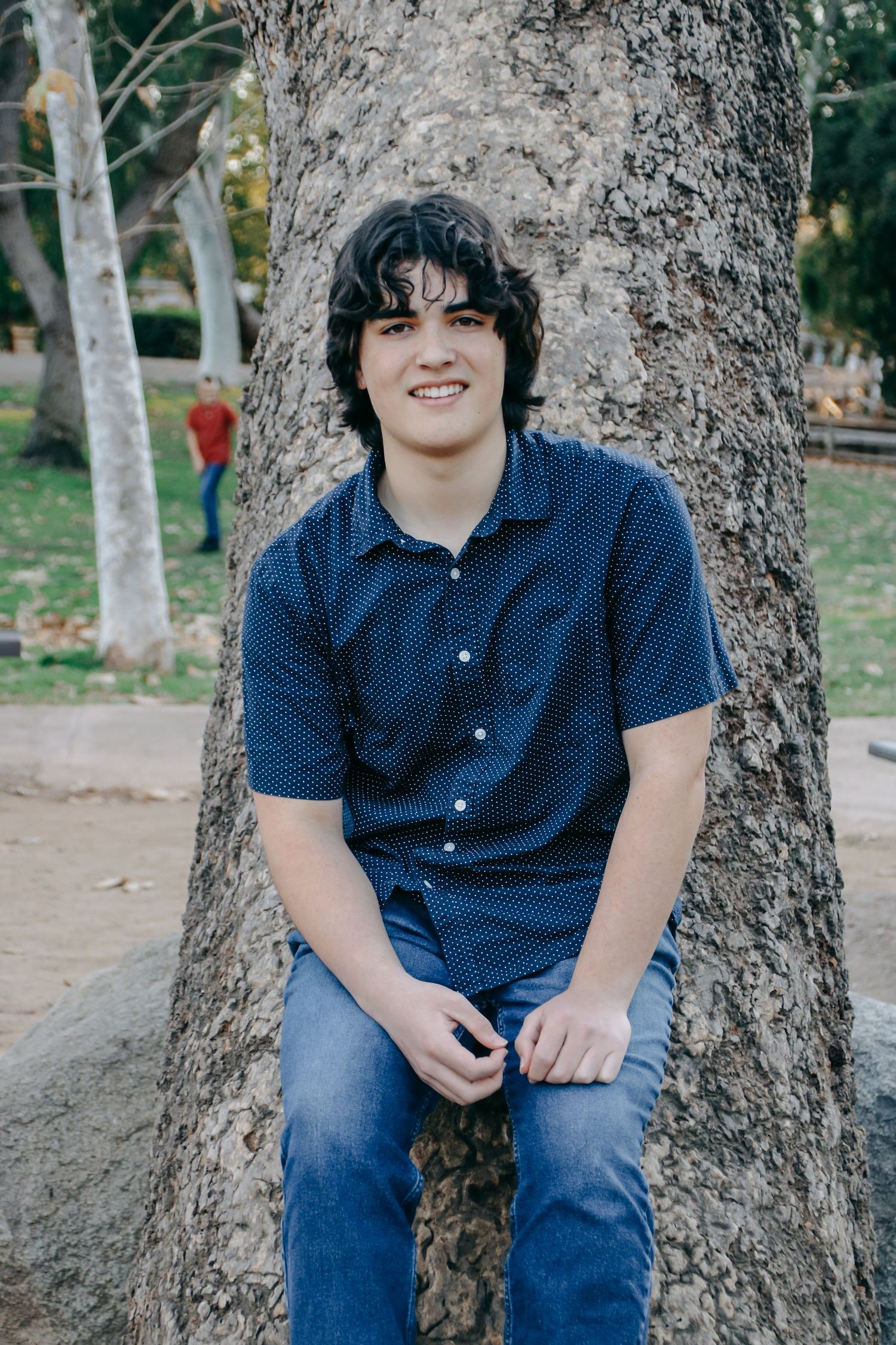 Someone relaxes against a tree trunk in an outdoor park setting wearing casual attire.