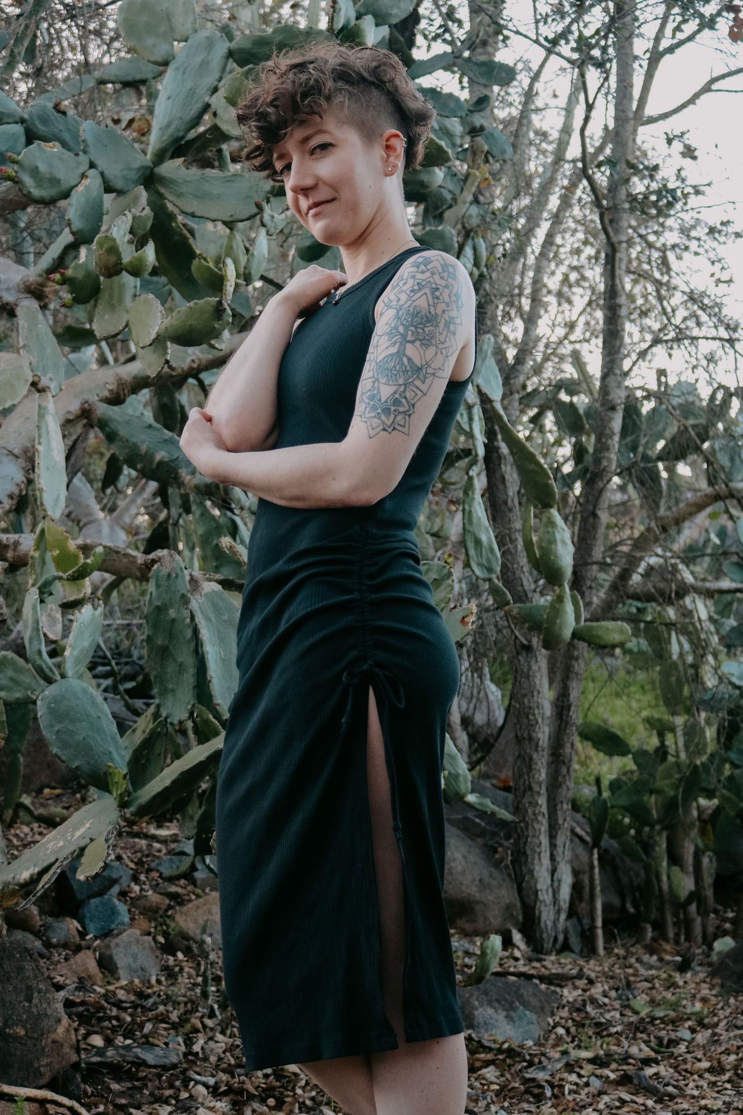 Person in sleeveless black dress strikes dramatic pose among desert vegetation and trees.