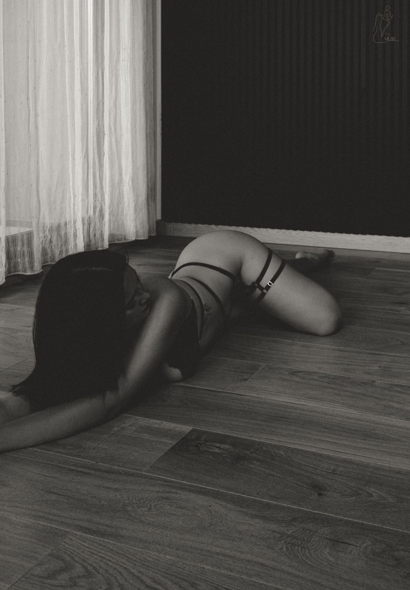 Black and white photo of a person lying on a hardwood floor in lingerie near a sheer curtain.