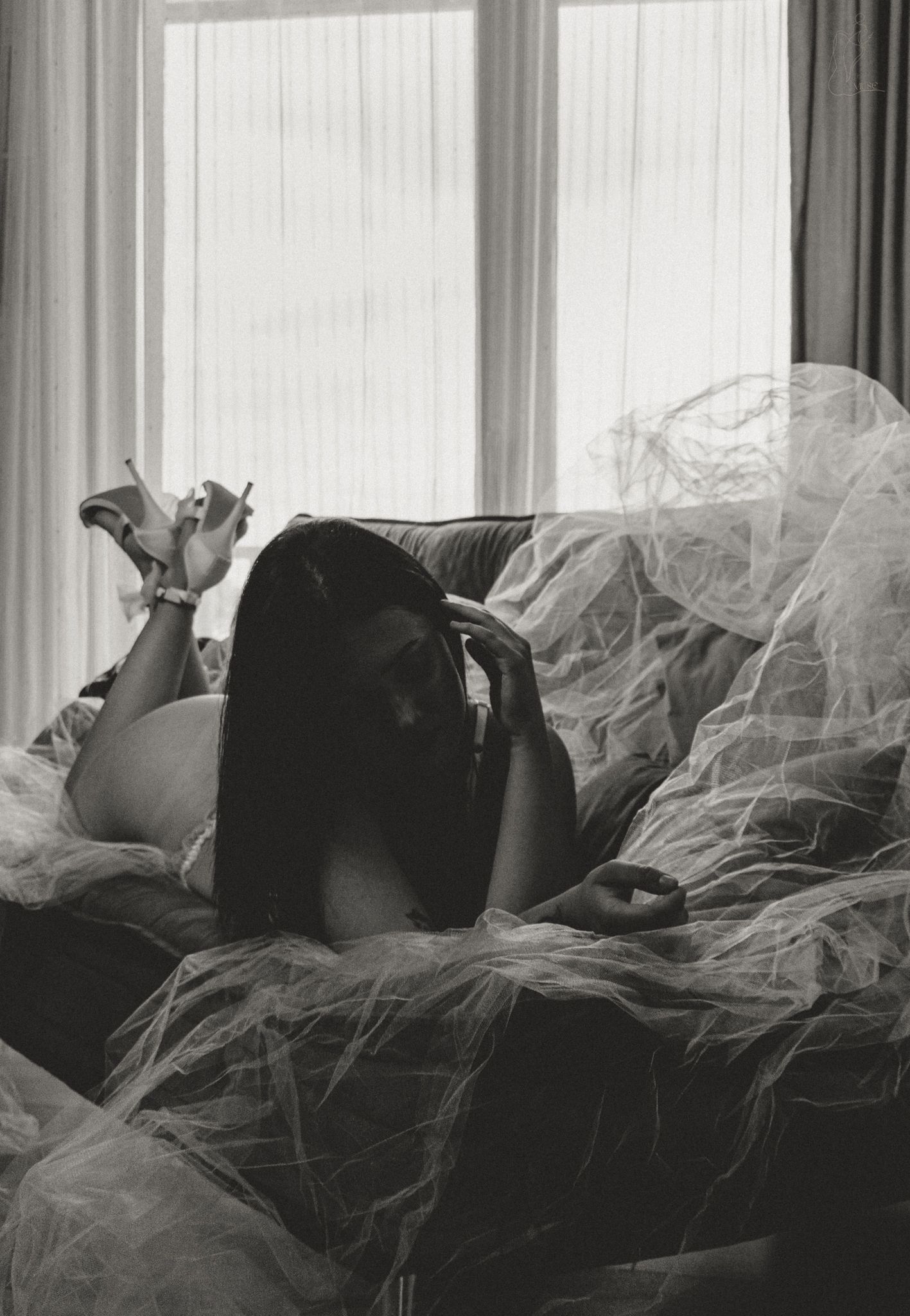 Black and white photo of a pregnant woman lying on a sofa draped in sheer tulle fabric near a bright window.