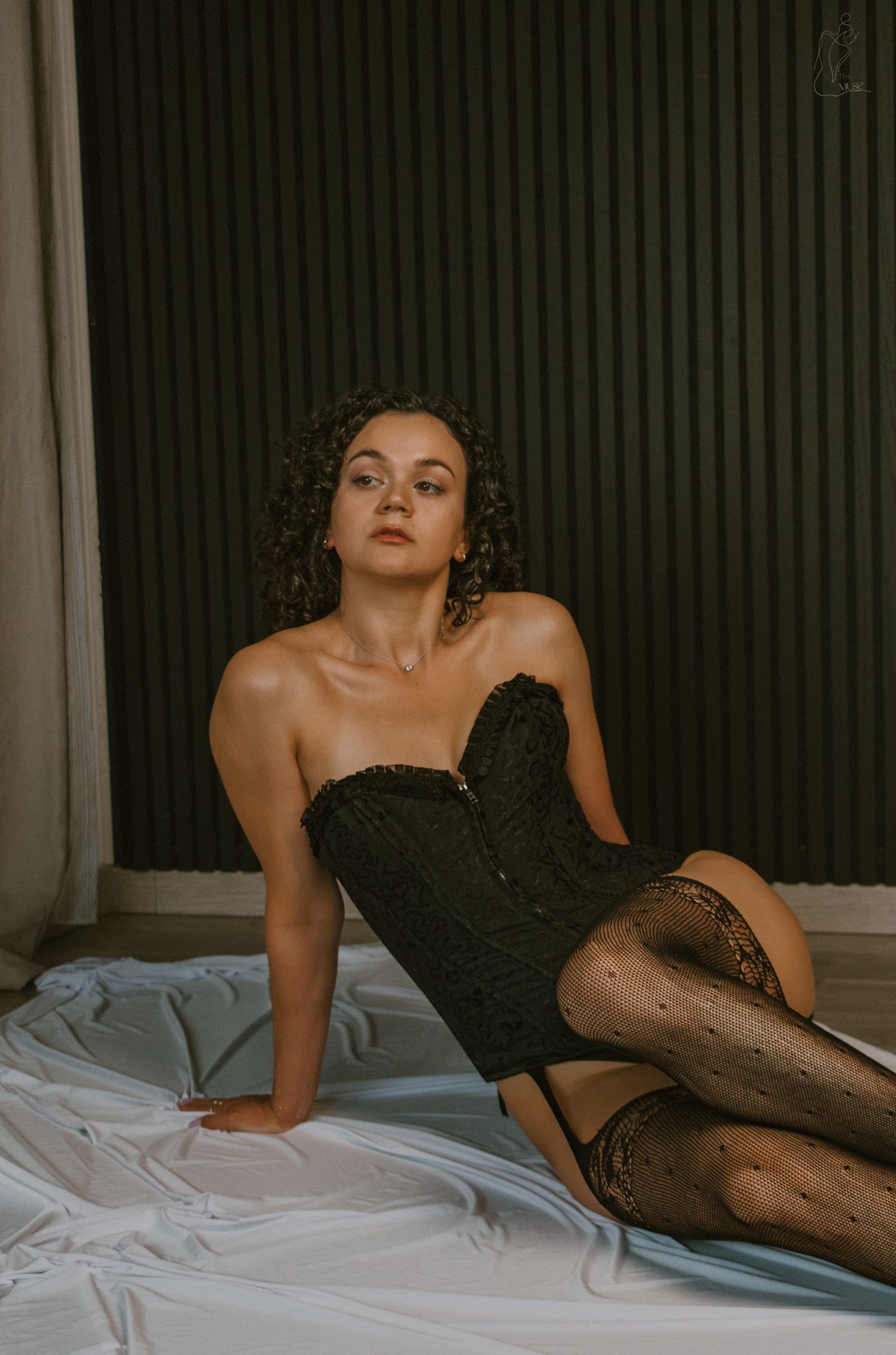 Woman with curly hair in a black strapless corset and polka dot stockings posing on white fabric.