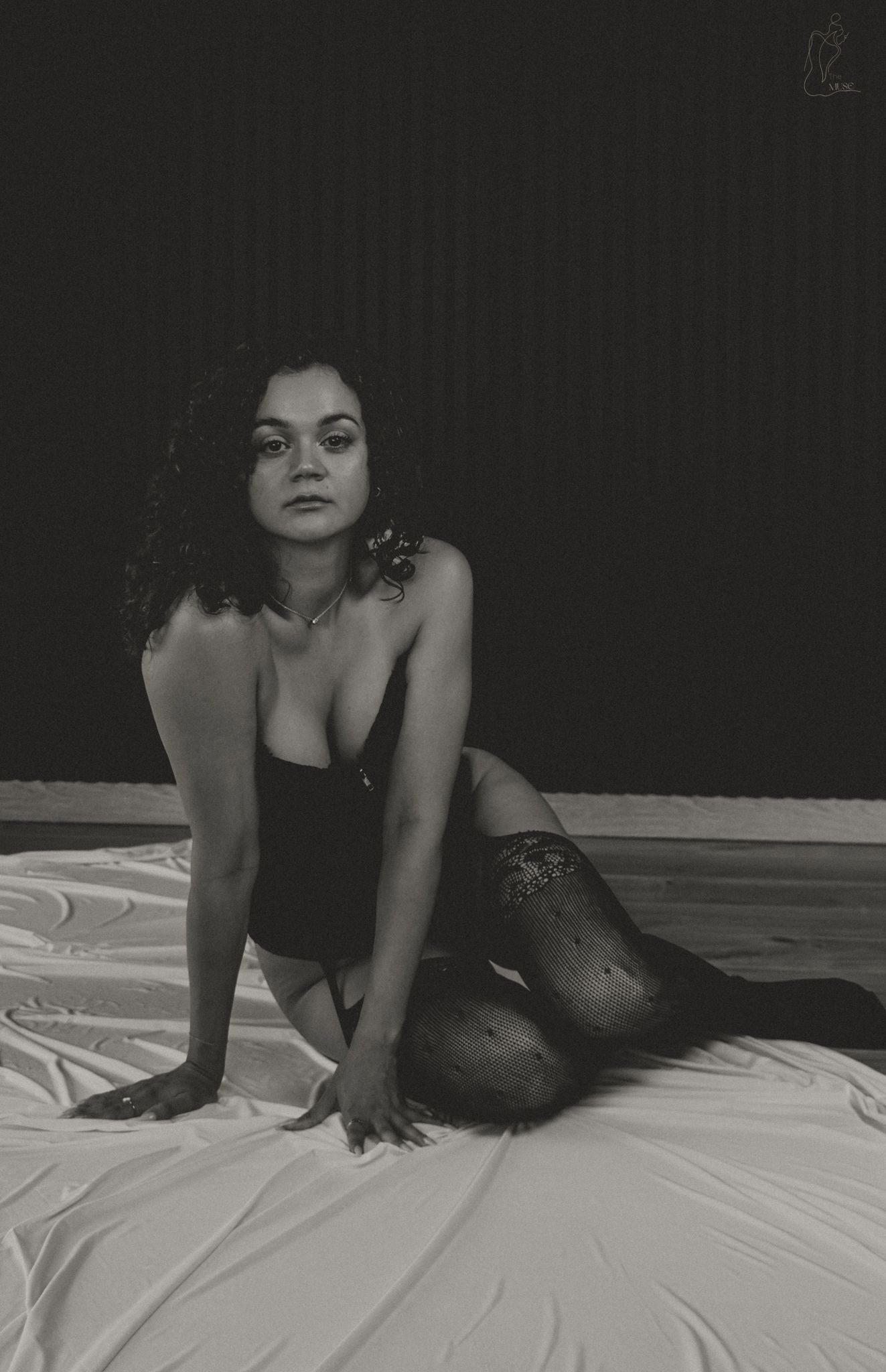 Black and white boudoir photo of woman in black bodysuit and lace stockings on white sheets.