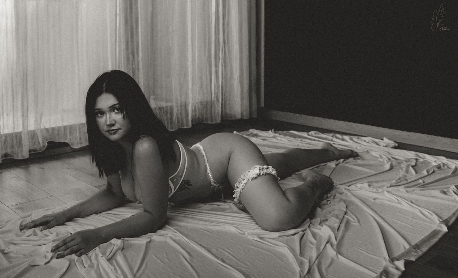 Black and white photo of a woman posing in lingerie on a bed near a window.