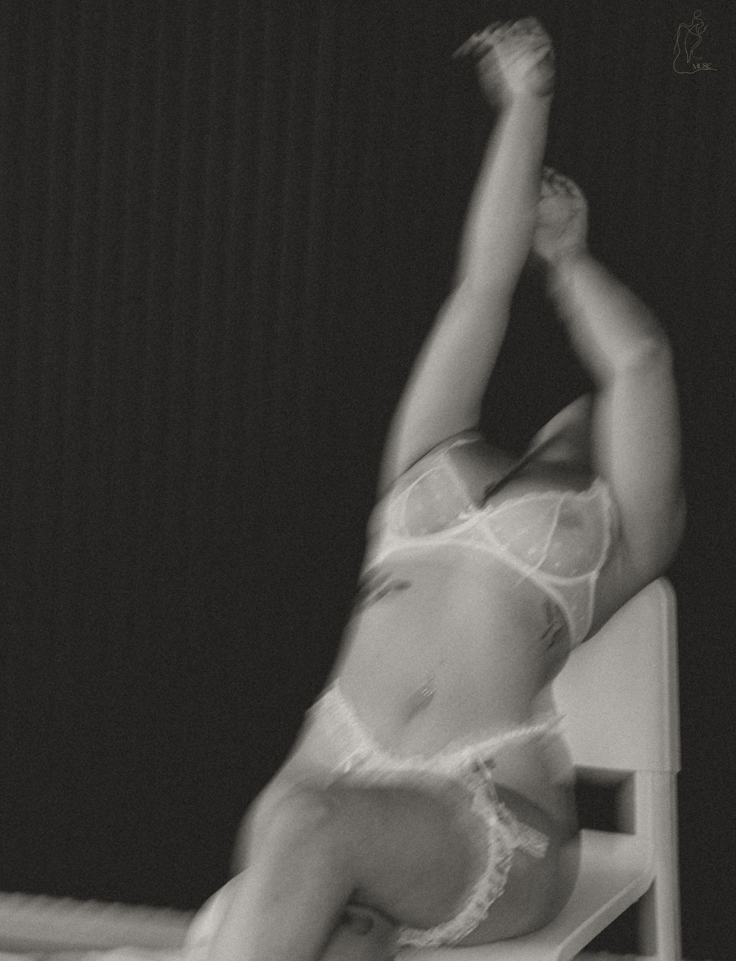 Black and white artistic photo of a woman in white lace lingerie posing on a chair with legs raised.