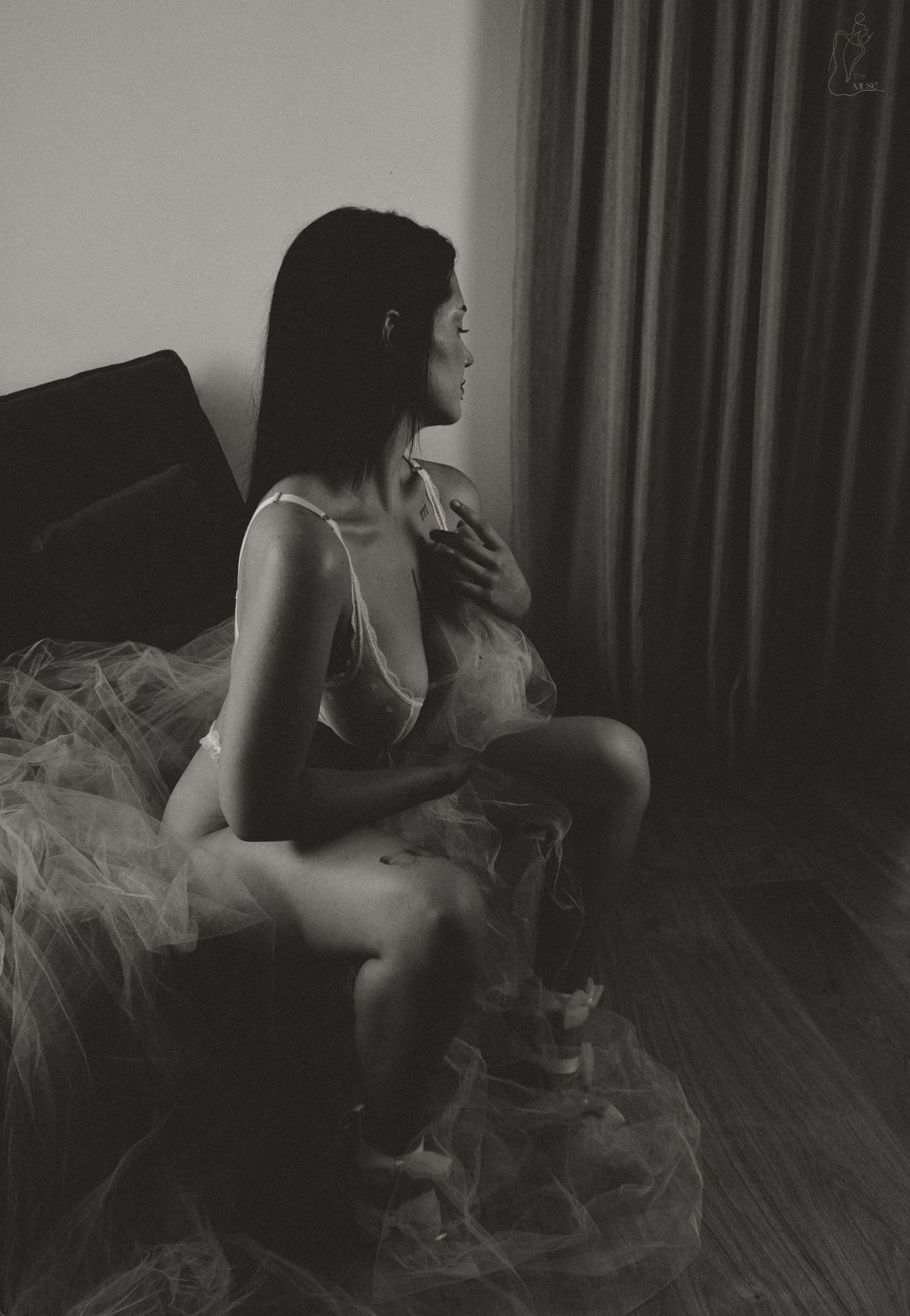 Black and white boudoir photo of a woman in lingerie sitting gracefully by a window with soft curtains.