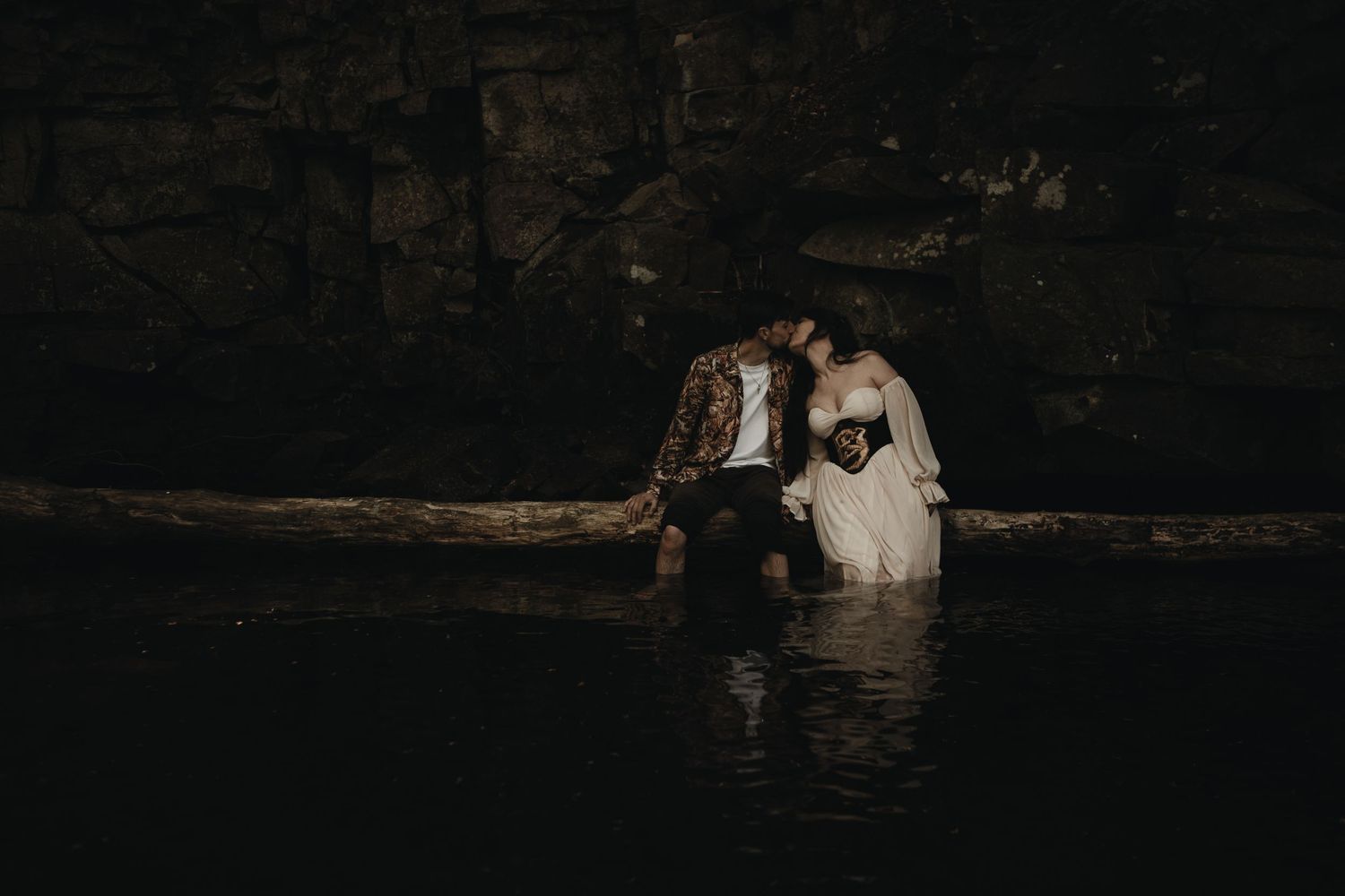 A couple shares a romantic kiss while sitting on a log over dark reflective water in a cave.