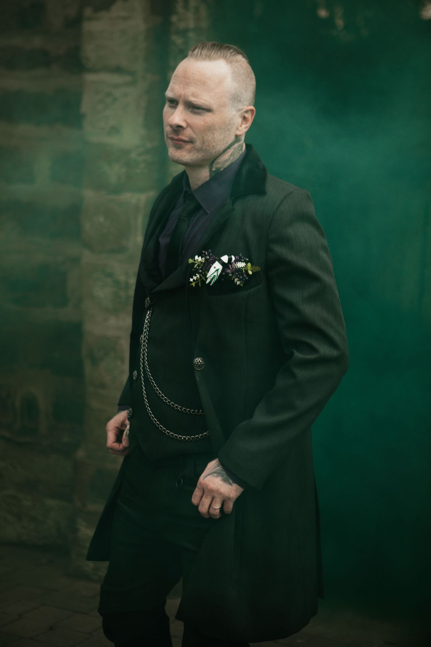 Man in black long coat with floral pocket square and chain, posing against stone wall with green smoke.