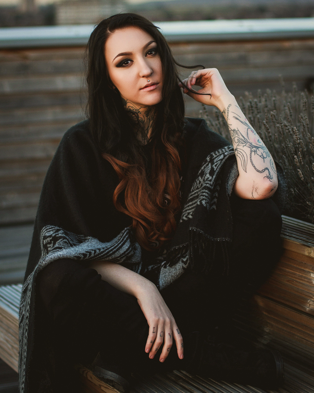 A tattooed woman with long dark hair poses in a black patterned wrap on a wooden bench outdoors.