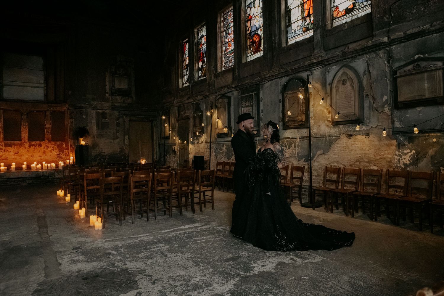 Couple in black gothic wedding attire embrace by candlelight in a dramatic stone chapel with stained glass windows.