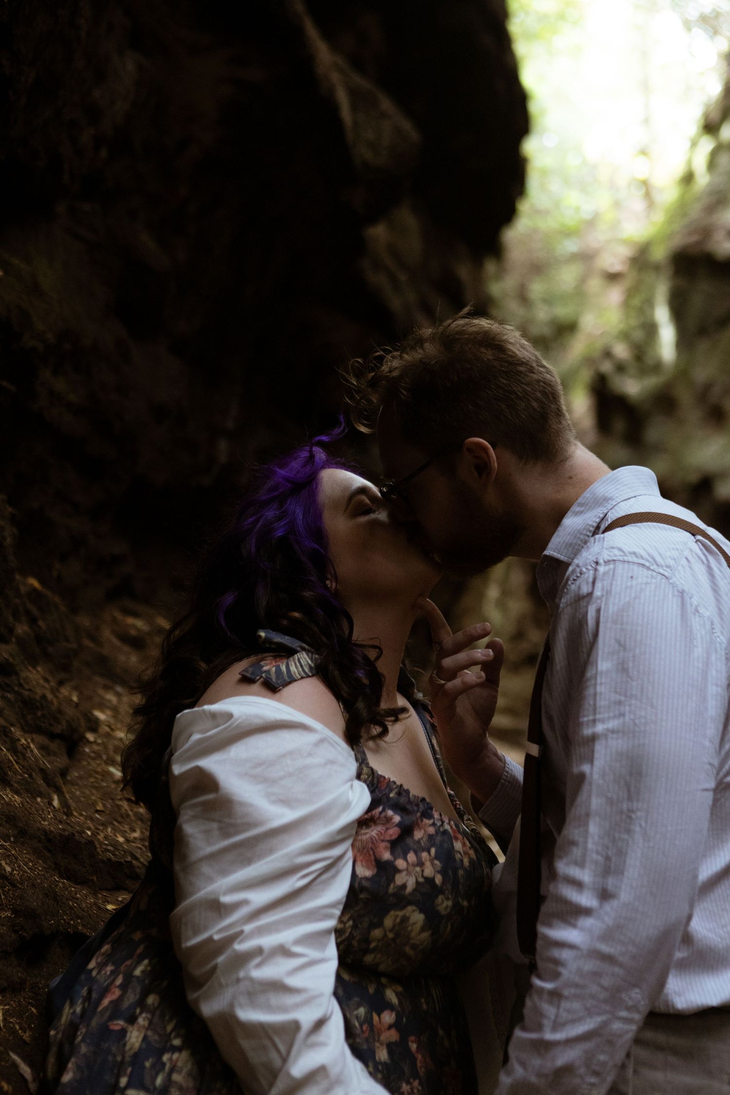 Couple sharing a romantic kiss in a dark forest canyon, woman with purple hair in floral dress, man in white shirt with suspenders.