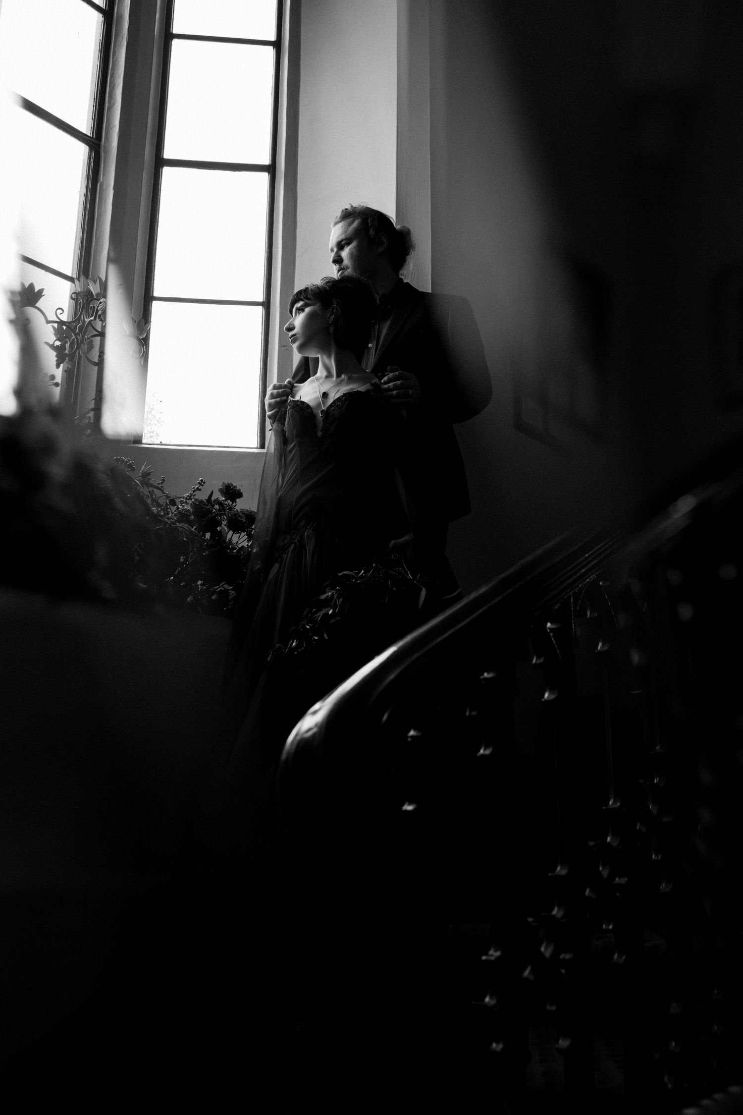 Black and white photo of a couple embracing by a large window on an elegant staircase in a classic interior.